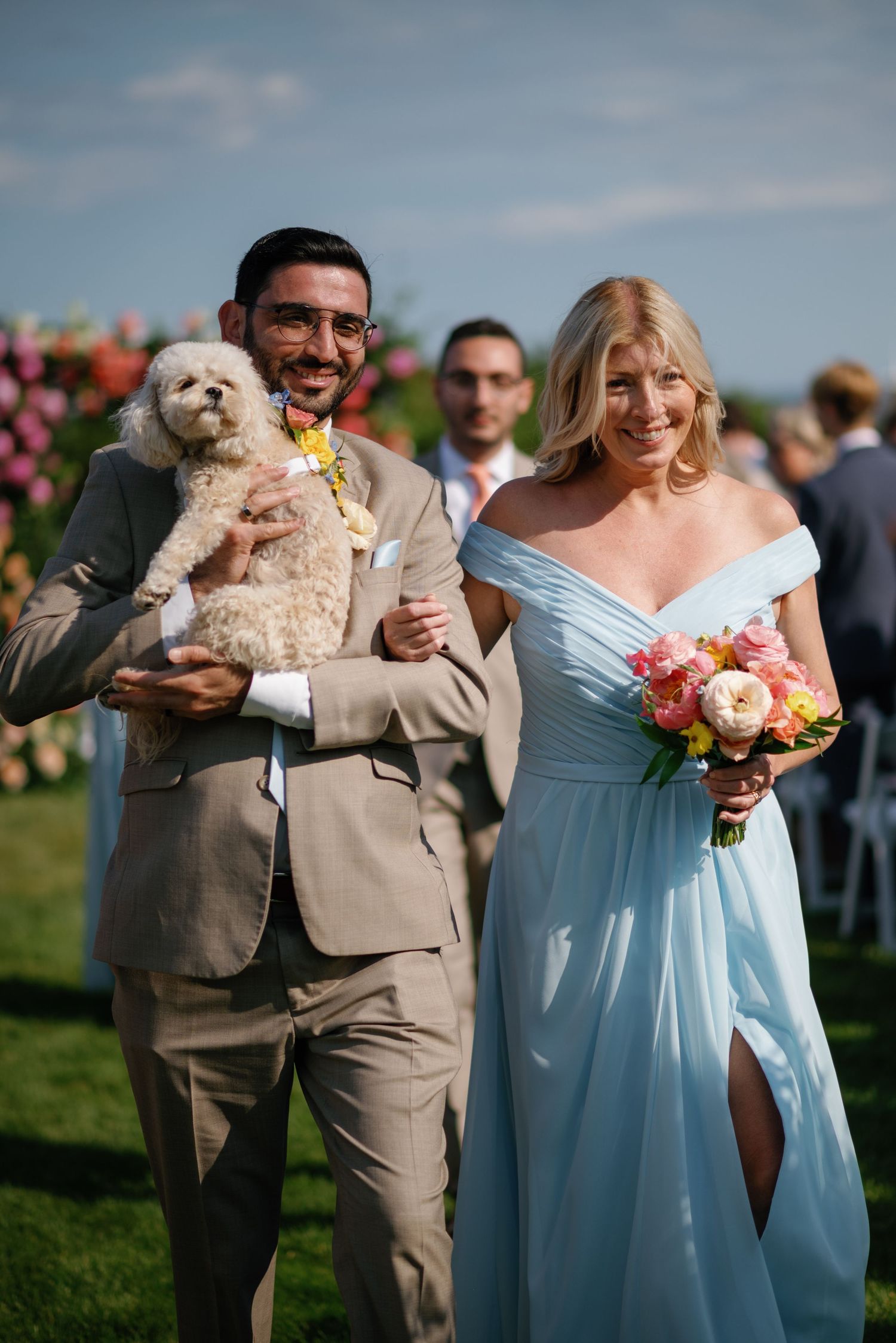 Wedding party members in light blue bridesmaid dress and tan suit pose outdoors with small white dog and floral bouquet.