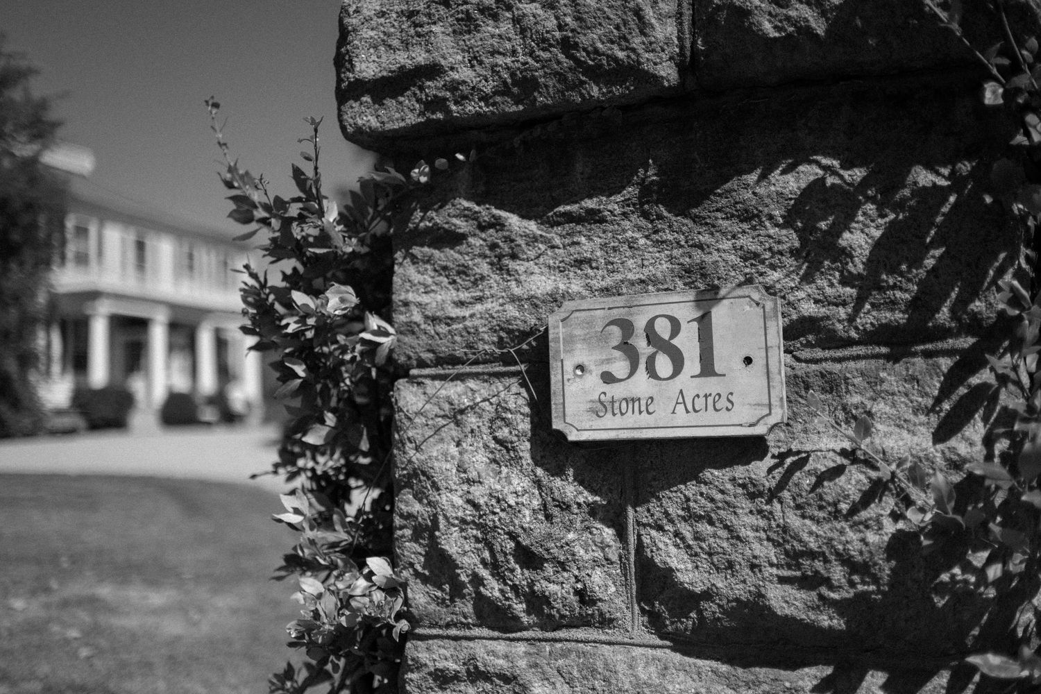Black and white photo of a stone pillar with address number 381 and Stone Acres sign, elegant estate in background.