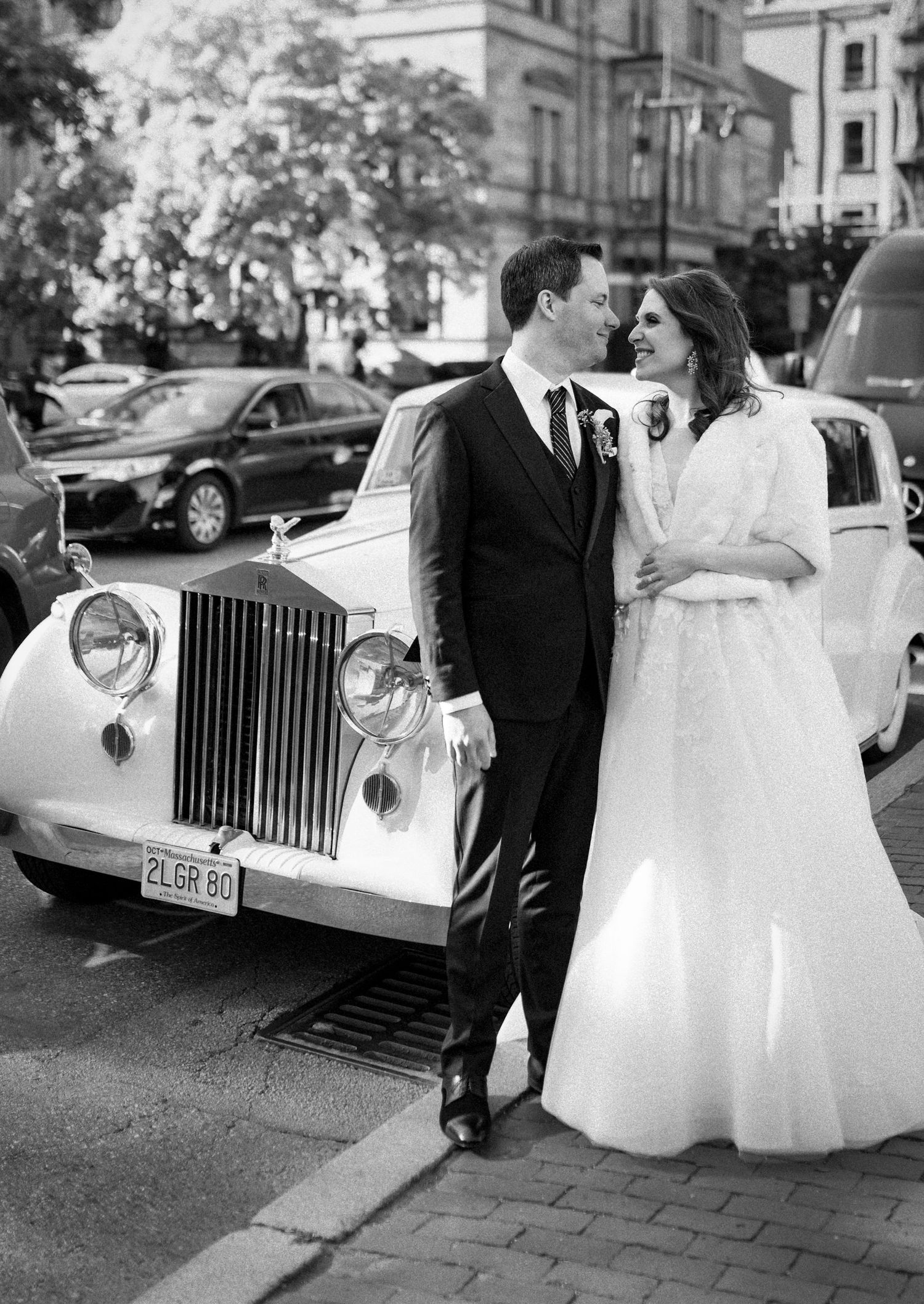 Black and white photo of a bride and groom posing beside a vintage Rolls-Royce on a city street.