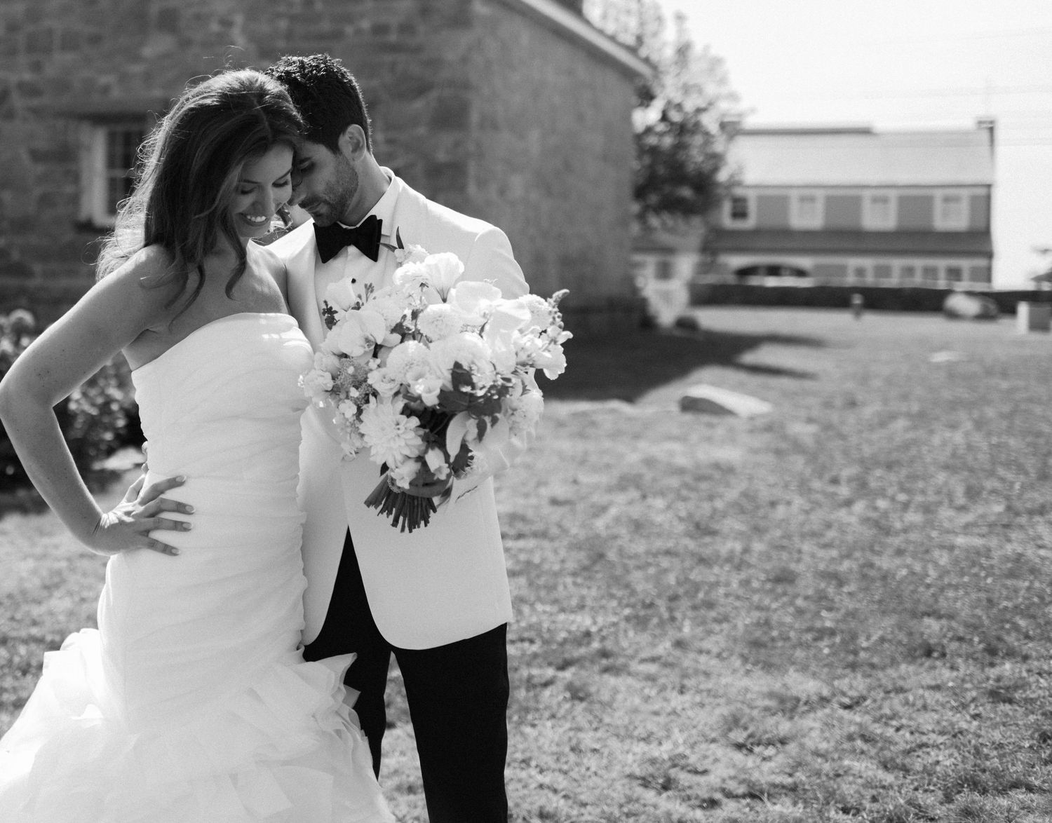 Black and white photo of a smiling bride holding white bouquet leaning into groom in white tuxedo outdoors.