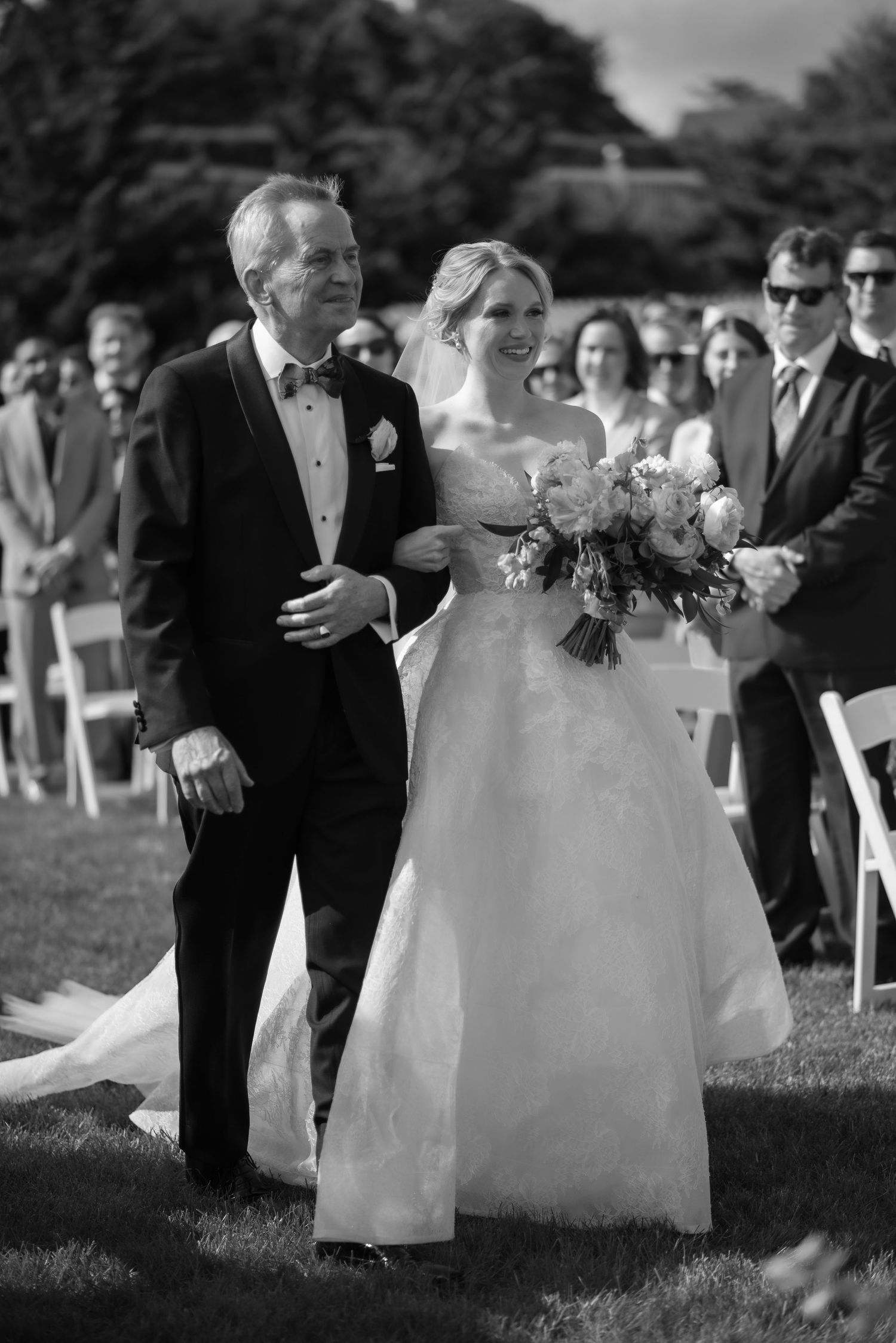 Black and white photo of a bride in a white gown holding flowers, walking down the aisle with a man in a tuxedo.
