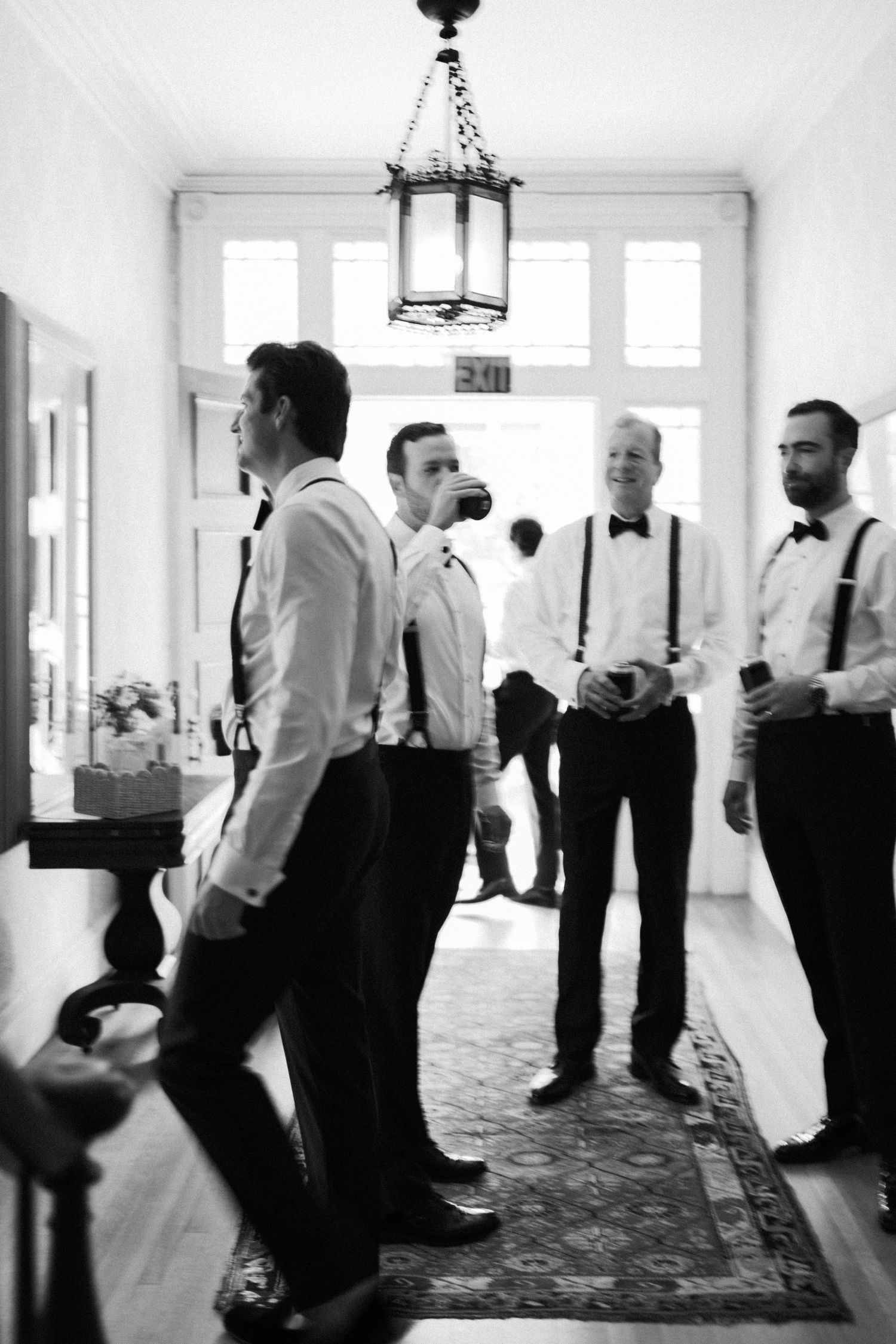 Groomsmen gather in a hallway with vintage lighting, wearing classic tuxedos and suspenders.