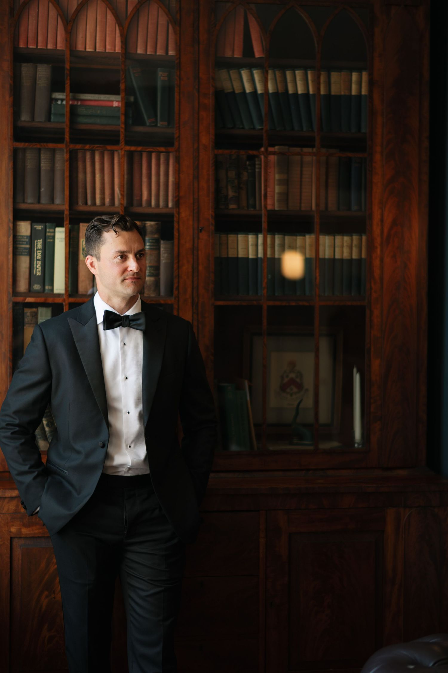 A man in a black tuxedo with bow tie stands confidently before a dark wooden bookcase filled with books.