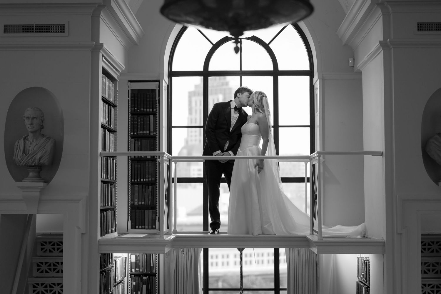 Black and white wedding photo of bride and groom kissing on balcony in elegant library with arched windows.