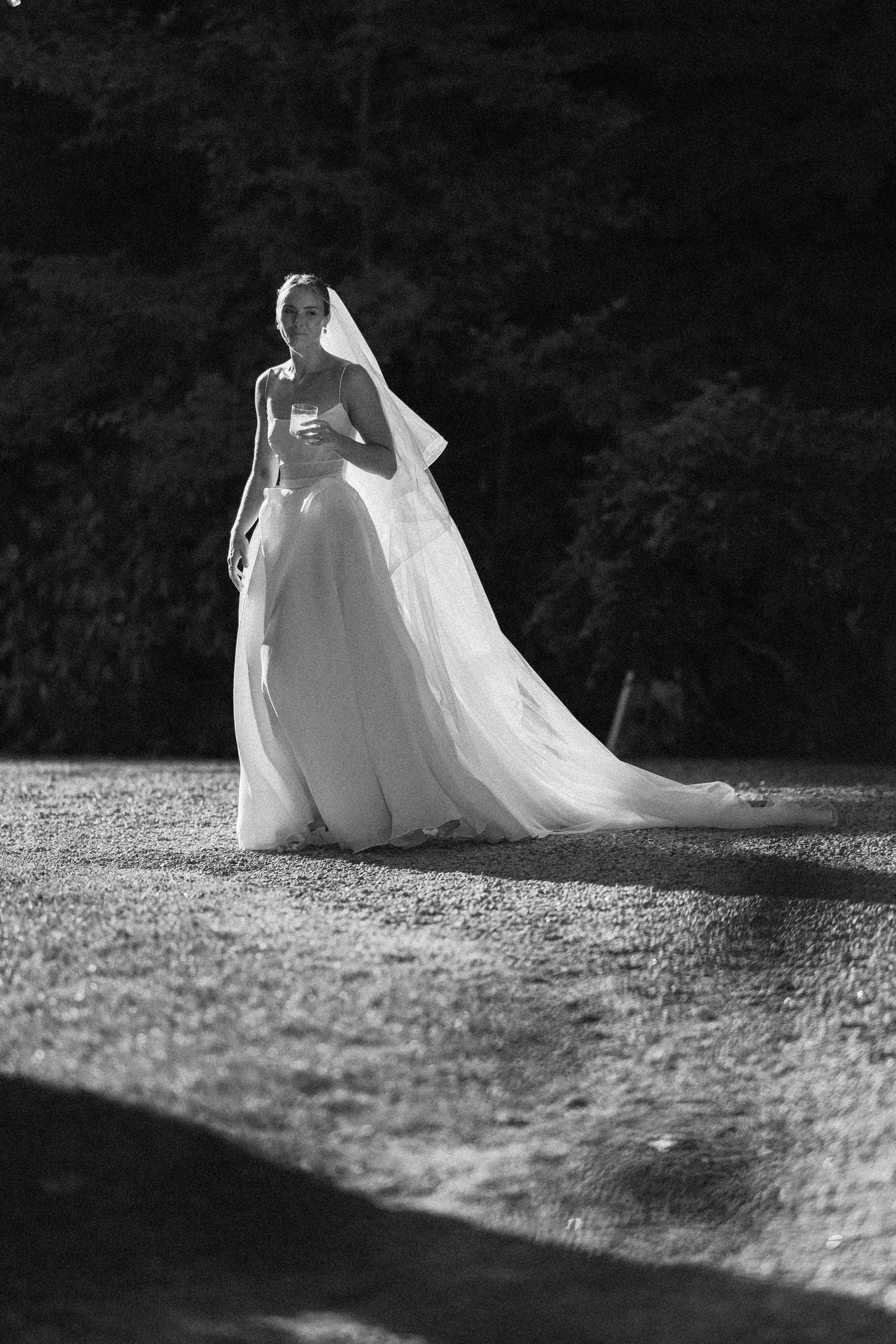 Black and white photo of a bride in a flowing white gown and veil standing outdoors on a sunlit path.