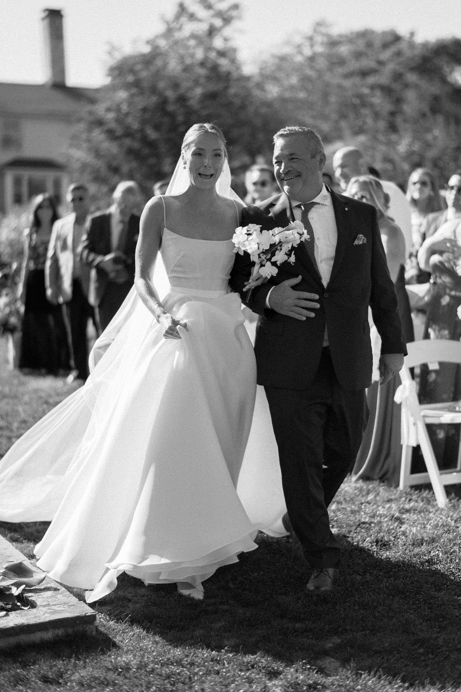 Black and white photo of a bride in a white gown walking down the aisle with an older man at an outdoor wedding.