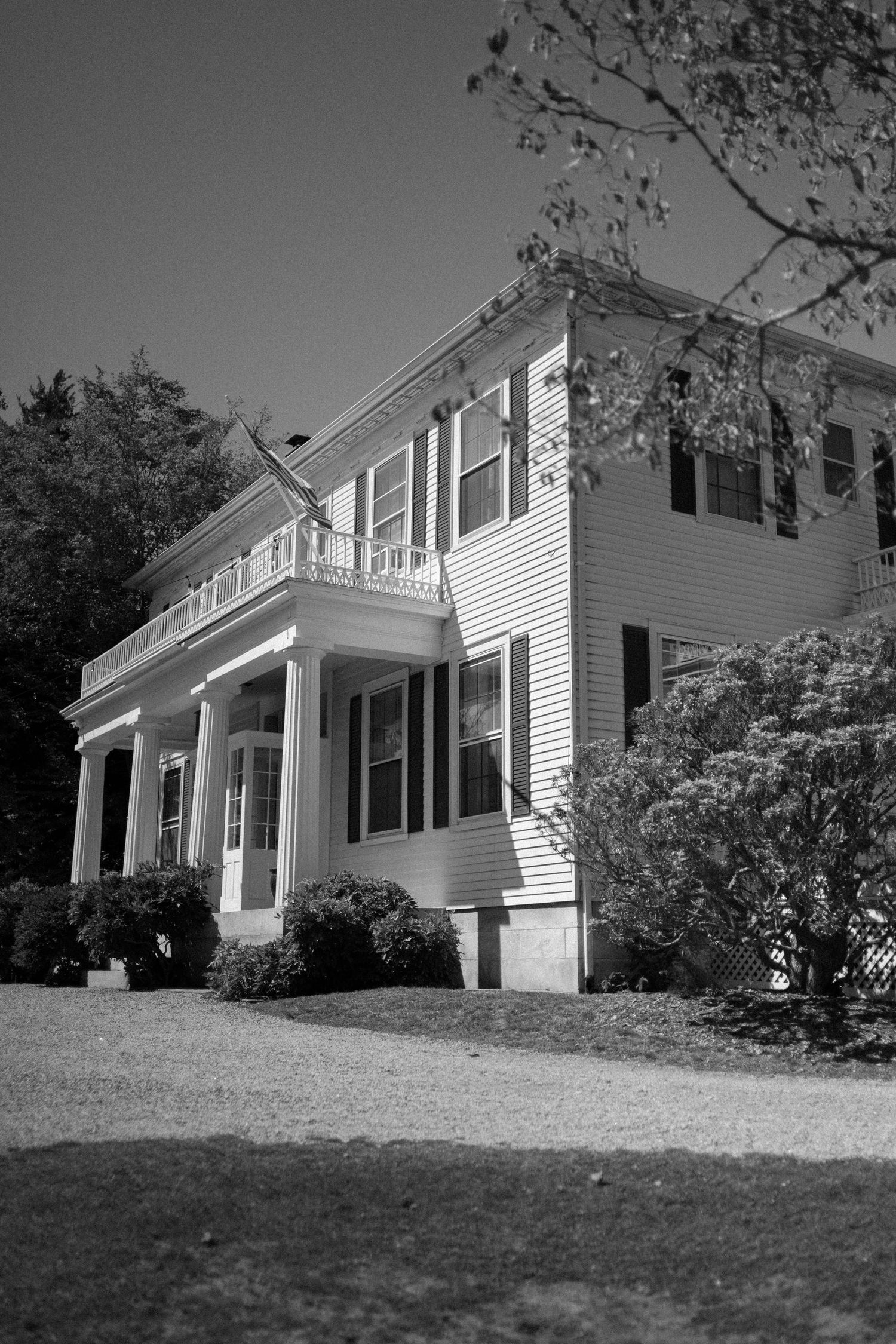 Black and white photo of a classic two-story colonial home with white columns, shuttered windows, and lush surrounding shrubs.