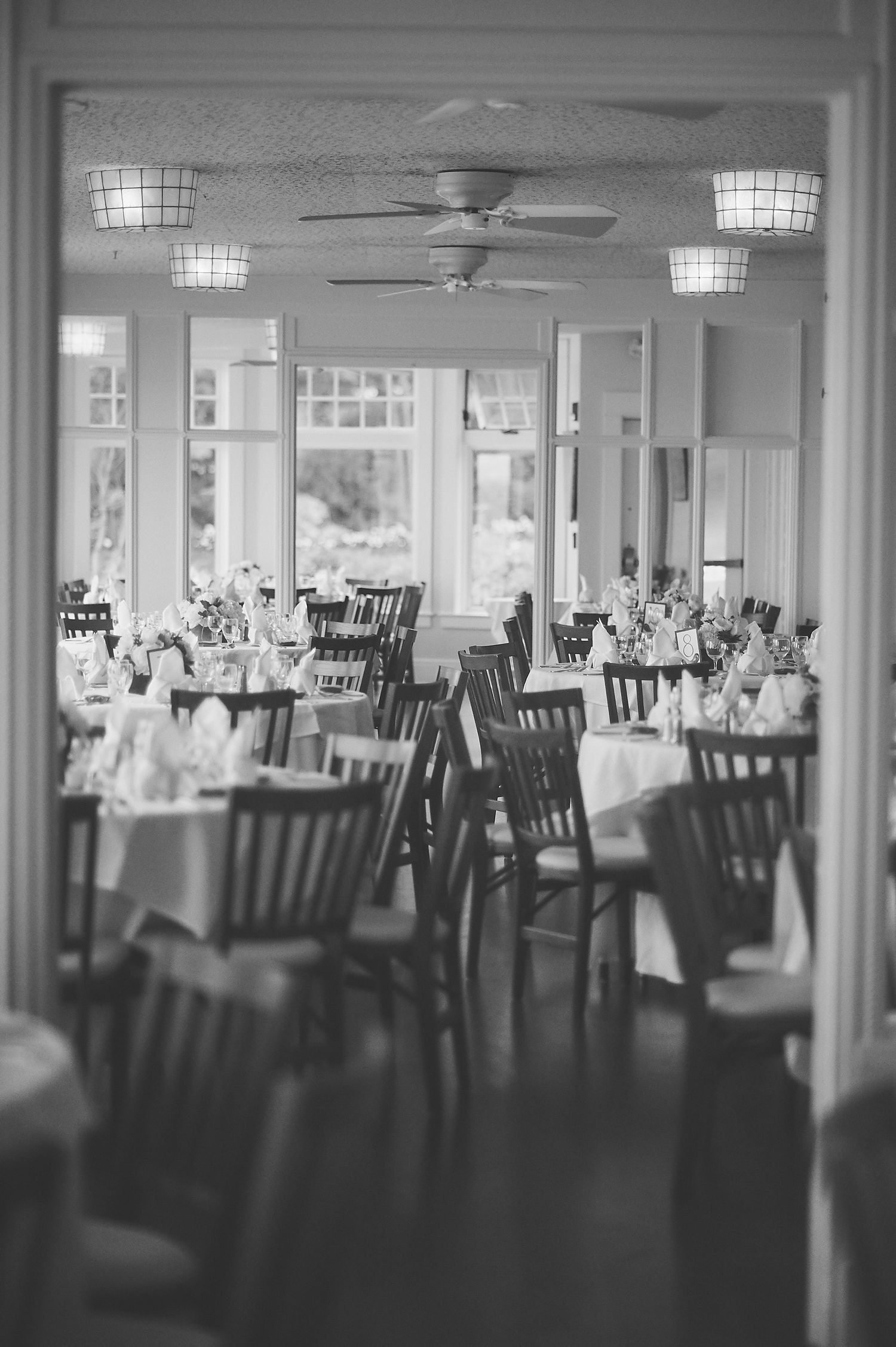 Black and white photo of empty wedding reception venue with wooden chairs and table settings.