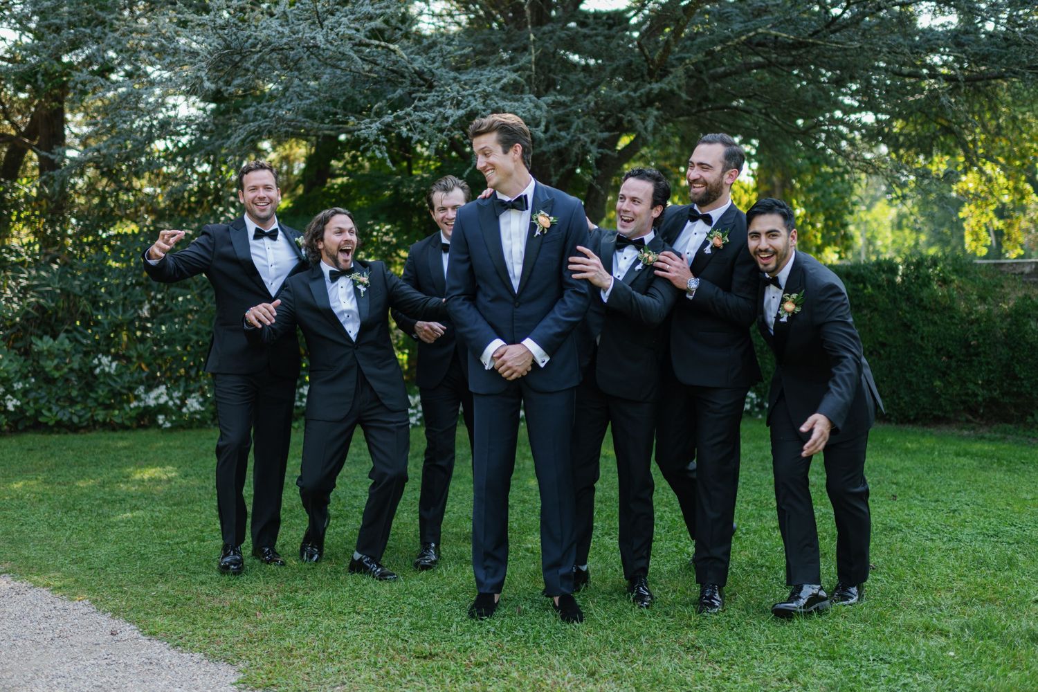 Groomsmen pose playfully on a garden path while wearing black tuxedos.