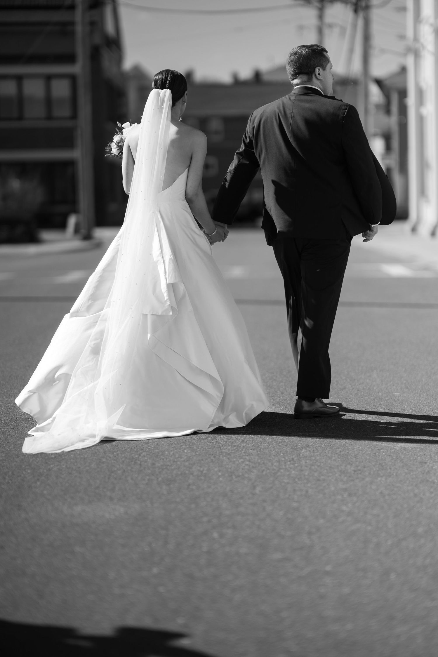 Black and white photo of a bride and groom walking hand in hand down a street, viewed from behind.