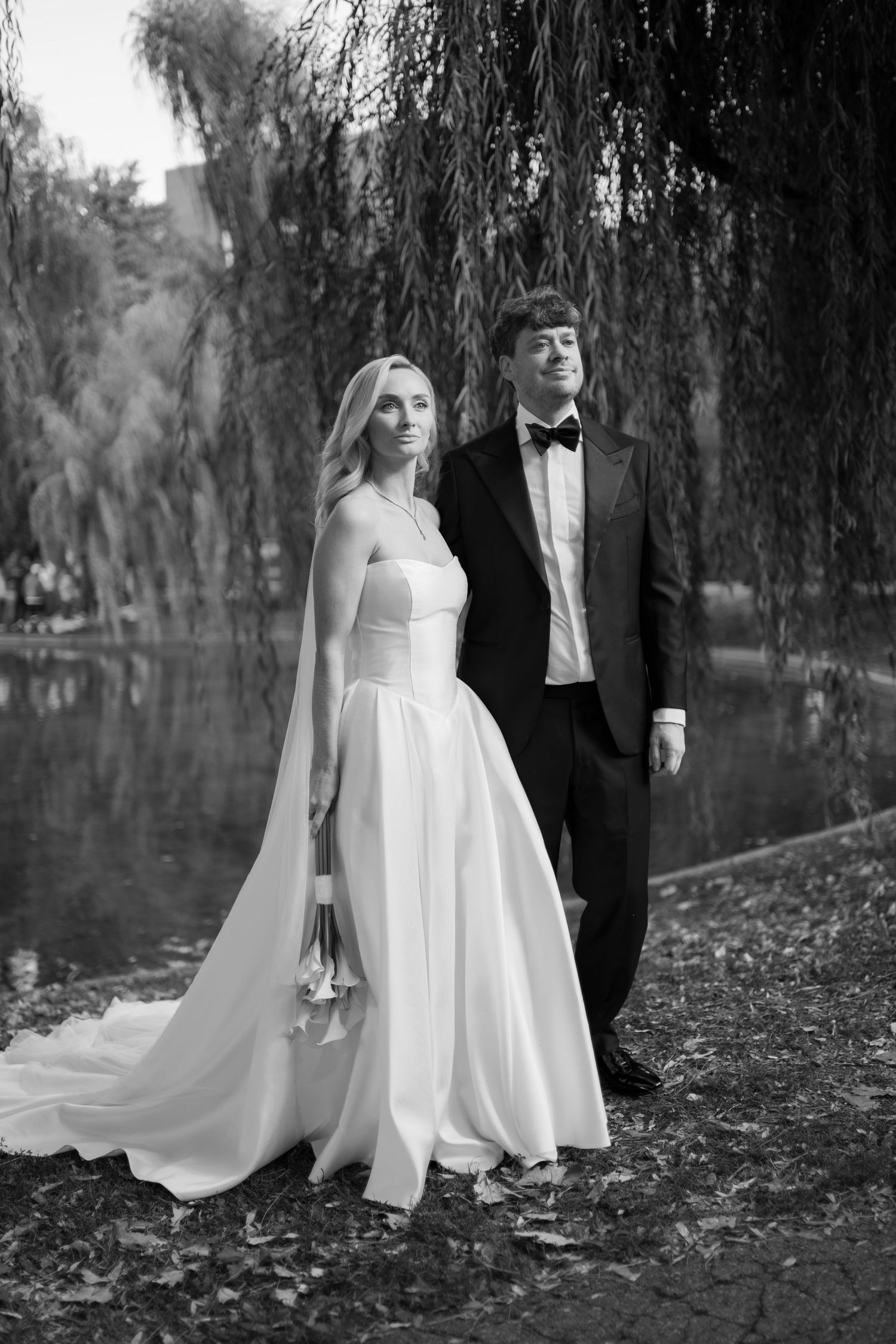 Black and white wedding photo of bride in white gown and groom in tuxedo standing by a pond under willow trees.