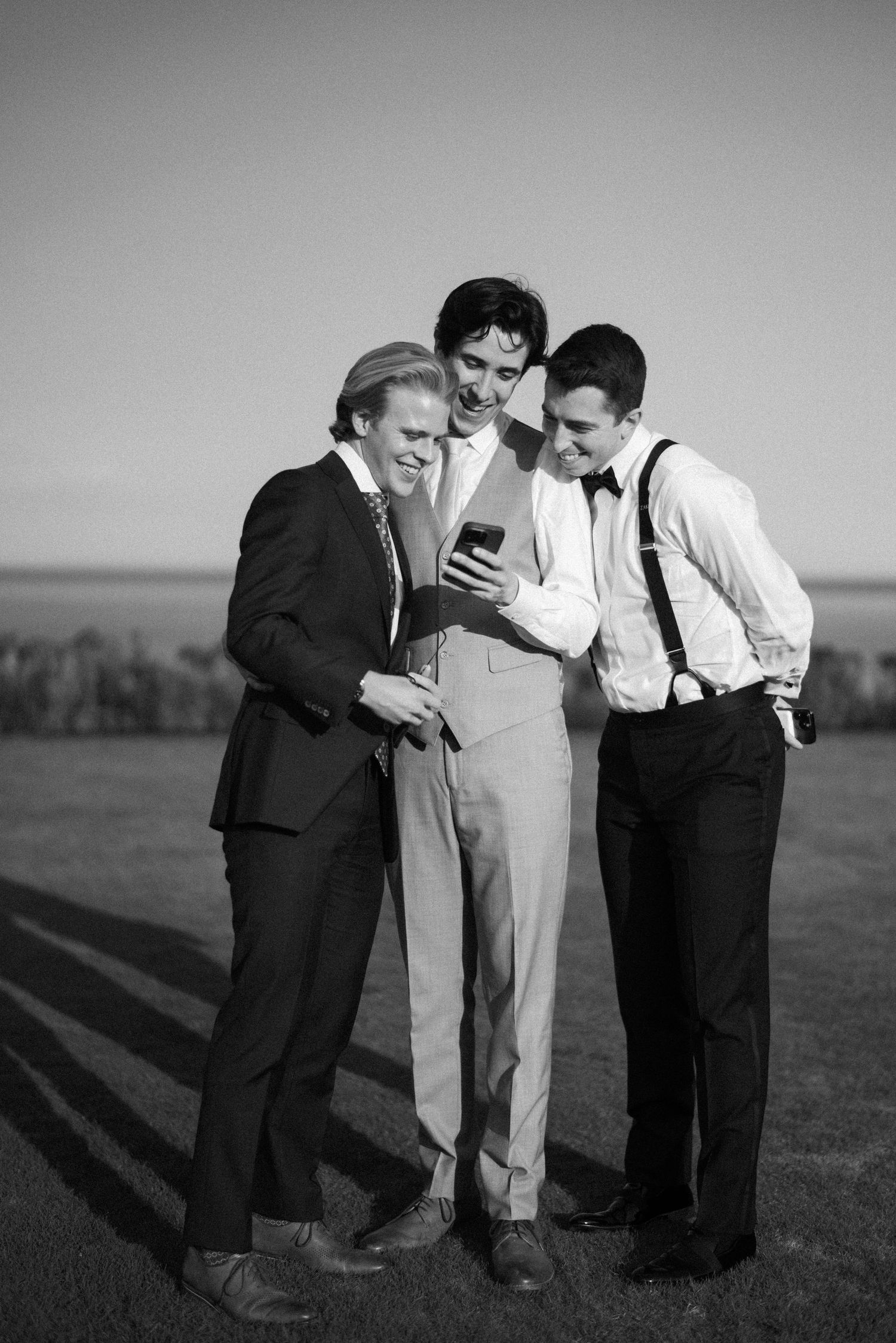 Three men in vintage attire huddle together looking down at something in a black and white photograph.