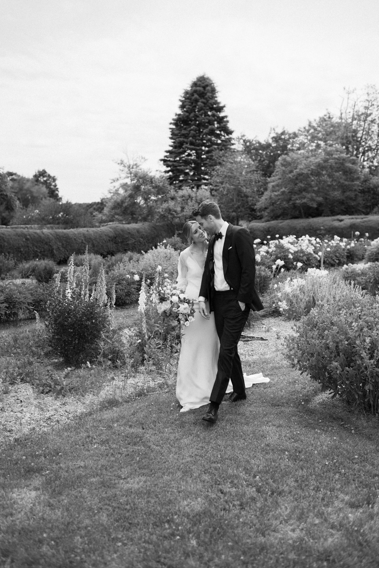 A romantic moment between newlyweds walking in a garden path surrounded by flowers and greenery.