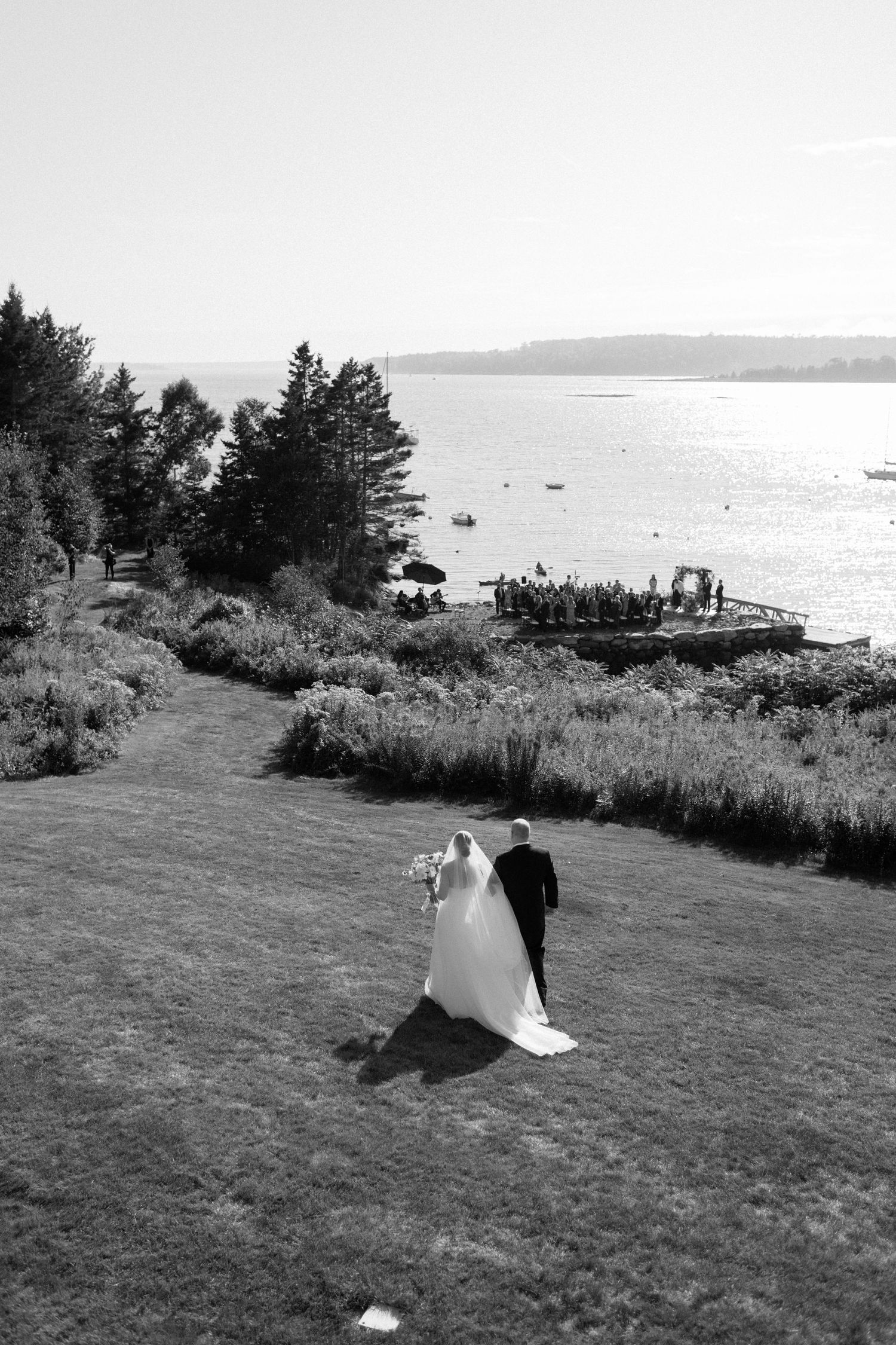 A couple walks on a grassy hill overlooking a scenic waterfront landscape in black and white.
