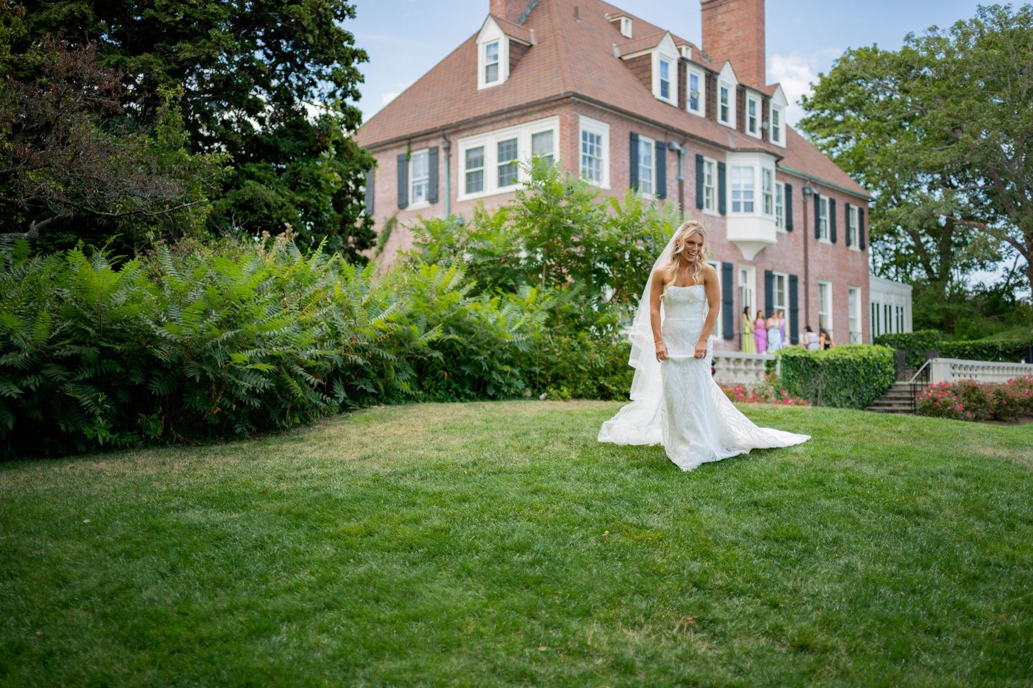 Bride in white wedding gown standing on lush green lawn in front of elegant brick colonial mansion.