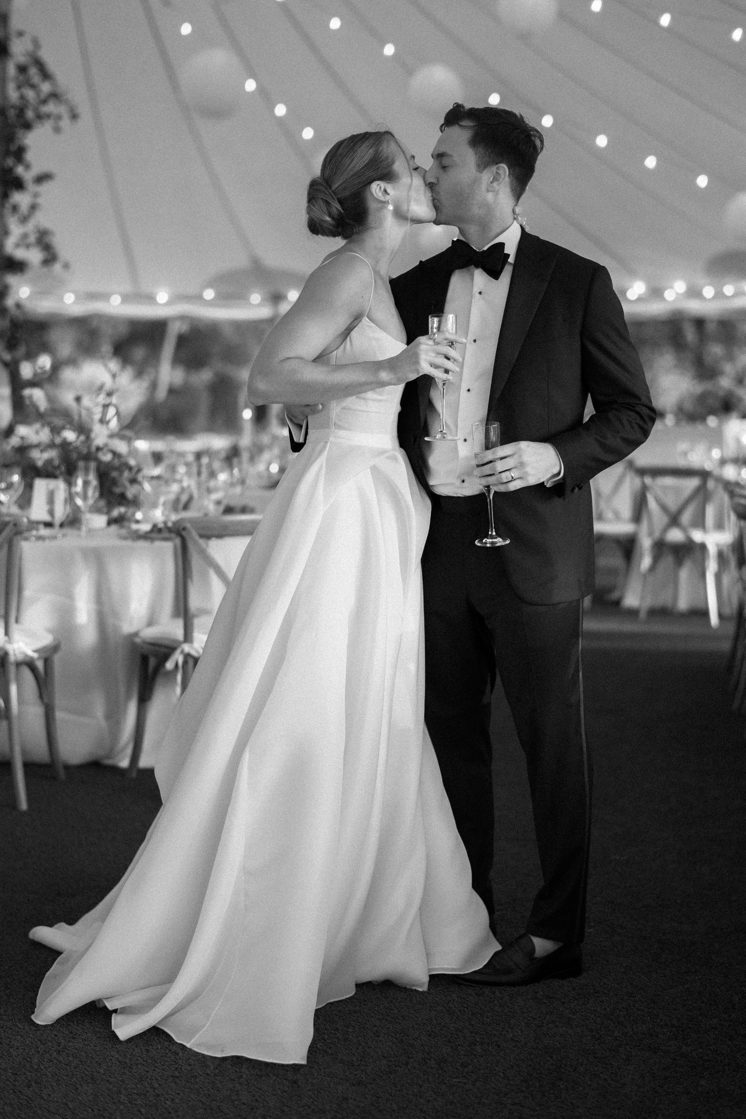 Black and white photo of a bride and groom sharing a kiss at their elegant tented wedding reception.