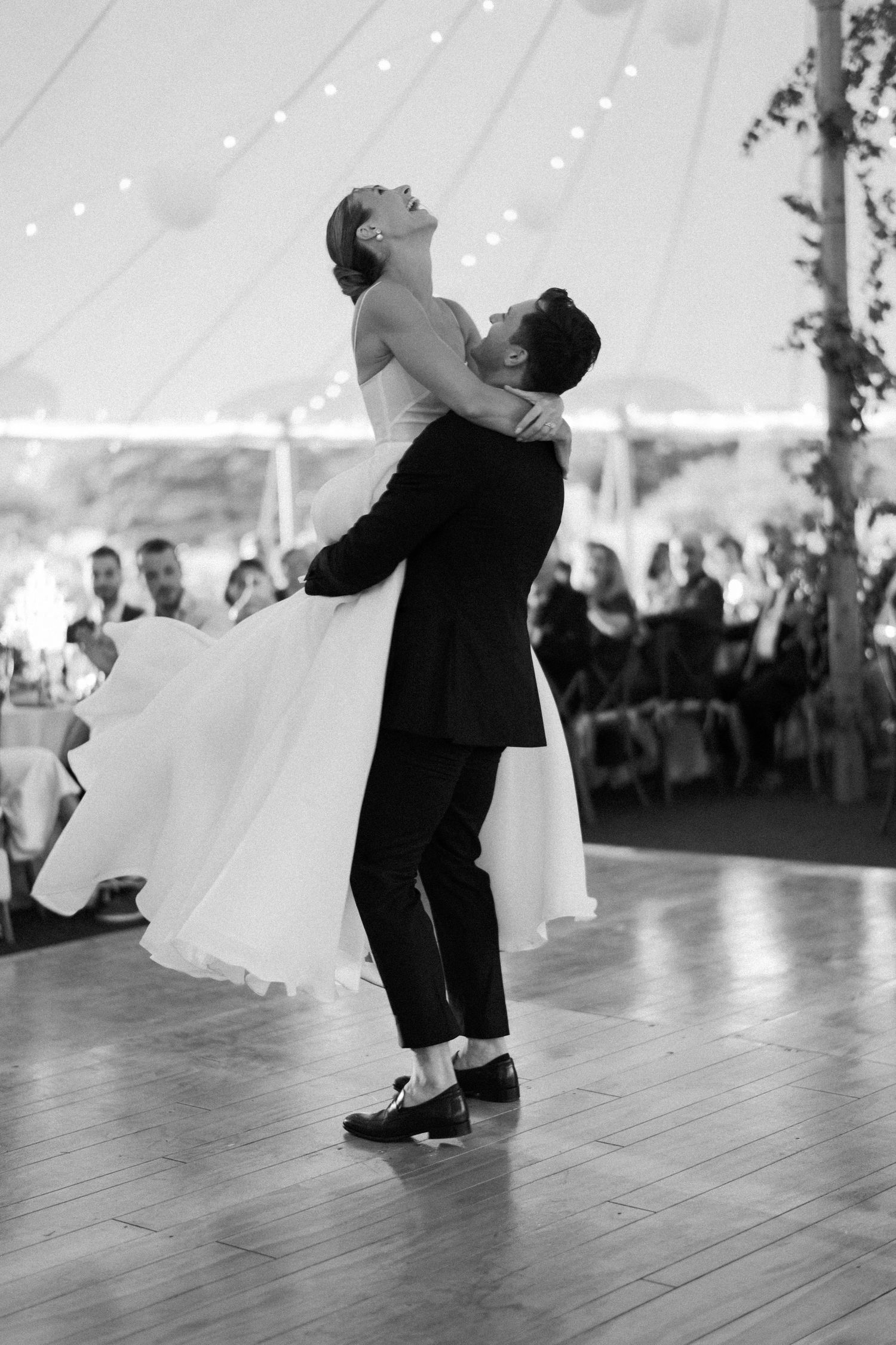 Groom lifts bride during first dance at elegant tented wedding reception in black and white photo.