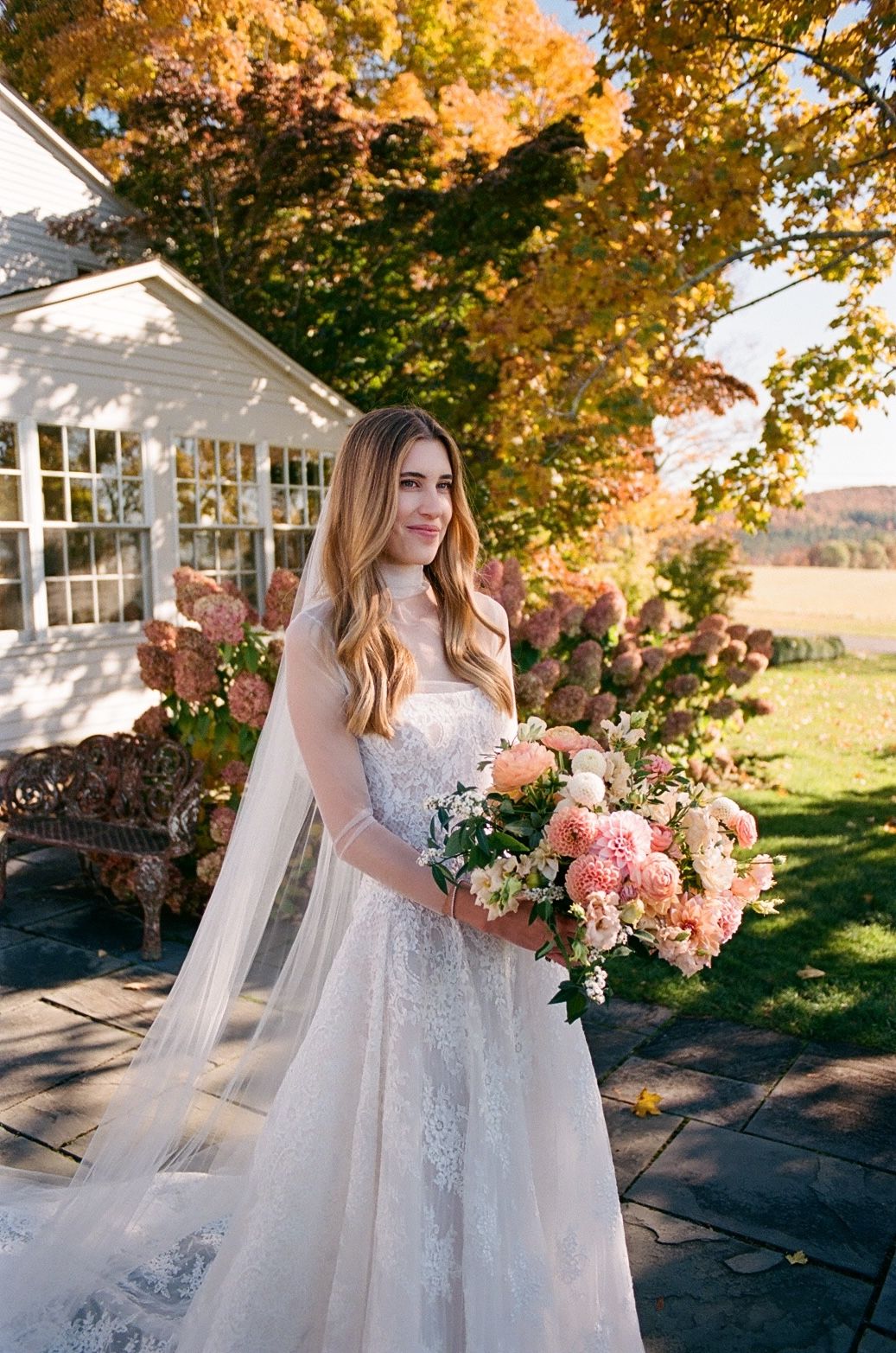 Bride in lace gown holds pink bouquet outdoors amid autumn foliage near a white building.
