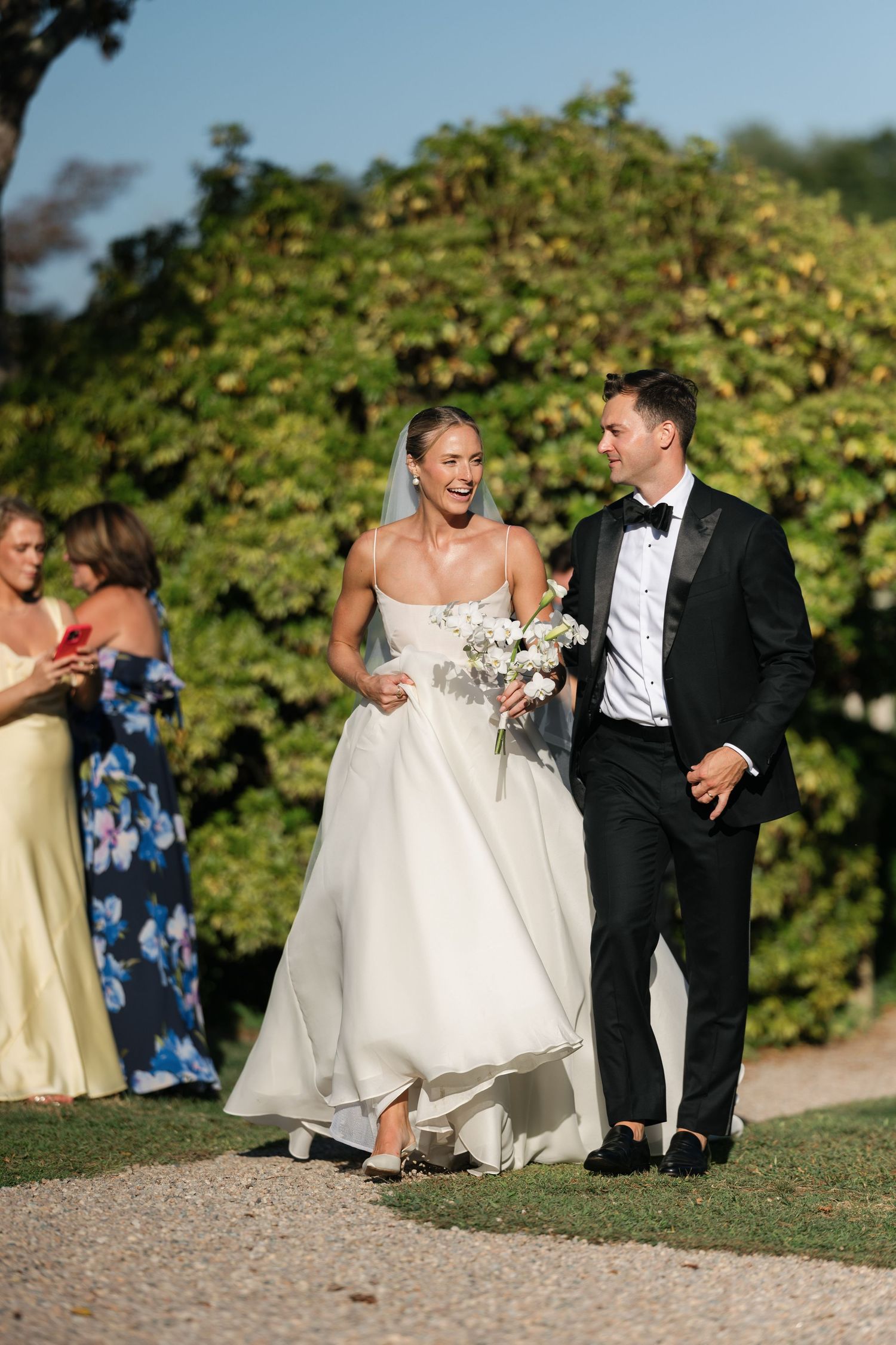 Smiling bride in white strapless gown and groom in black tuxedo walk together outdoors after wedding ceremony.