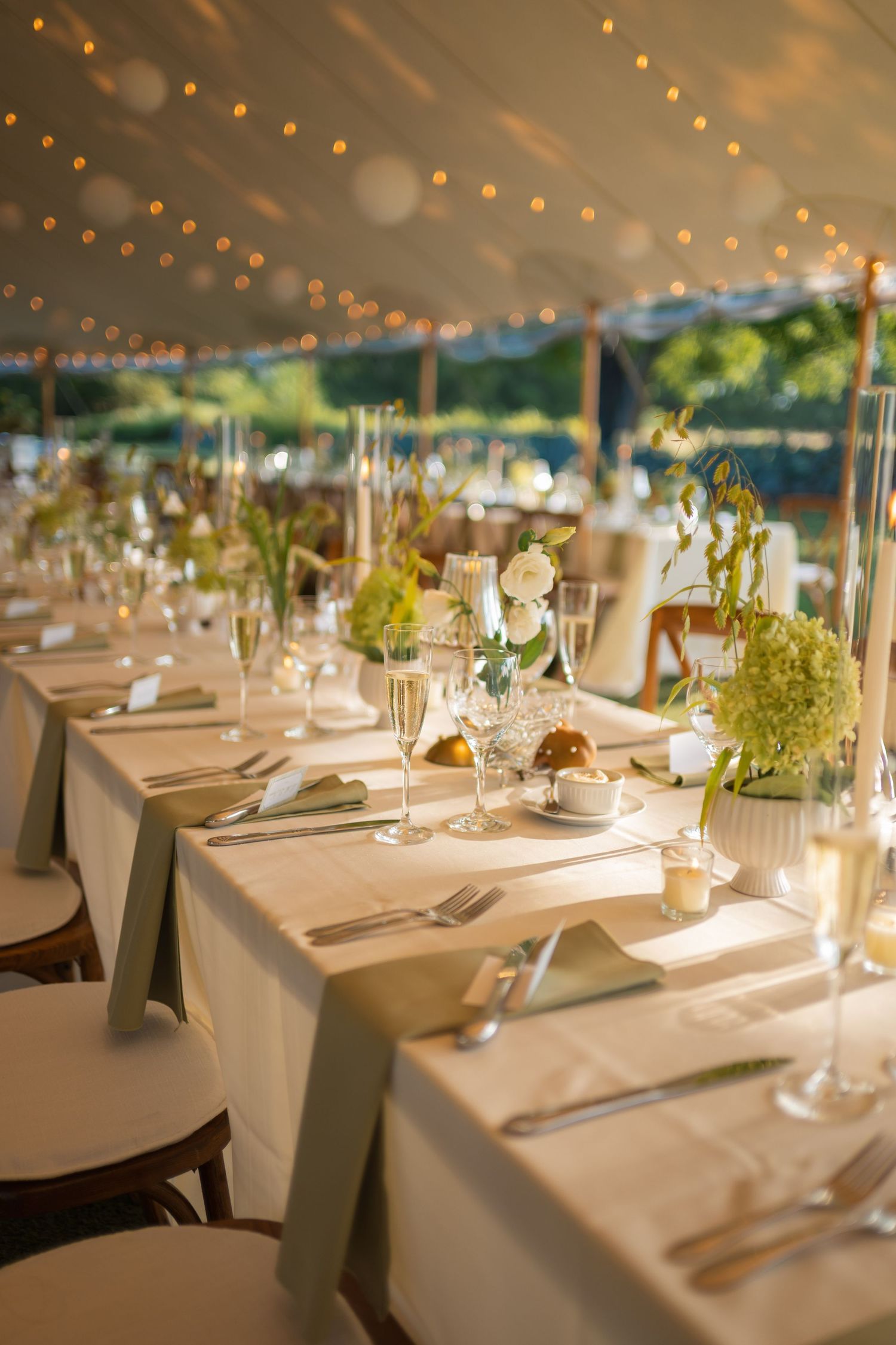 Elegant wedding reception tent with long tables, sage napkins, floral centerpieces, champagne flutes, and string lights overhead.