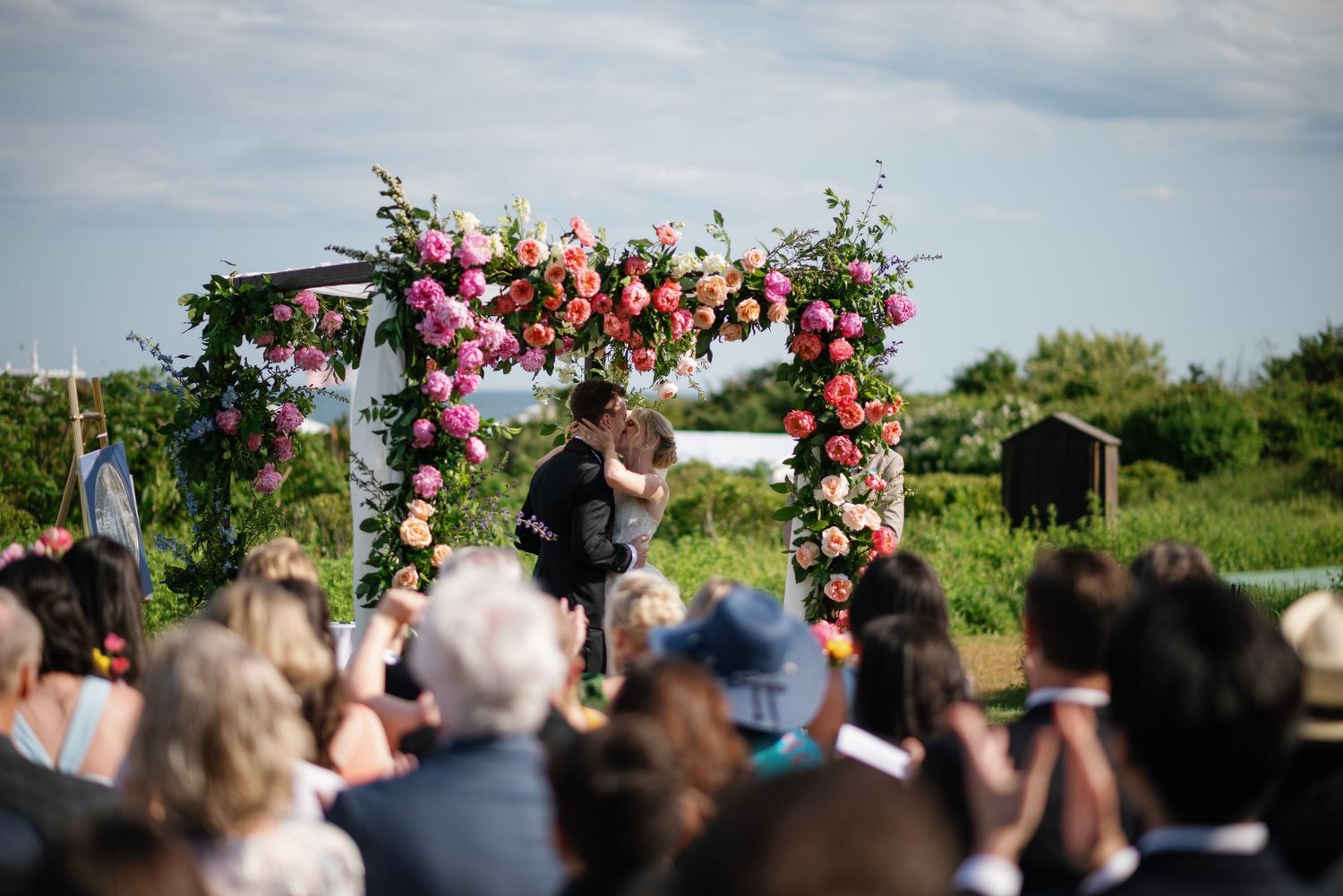 Wedding ceremony guests gather outdoors under blue skies with floral decorations and natural landscape in background.