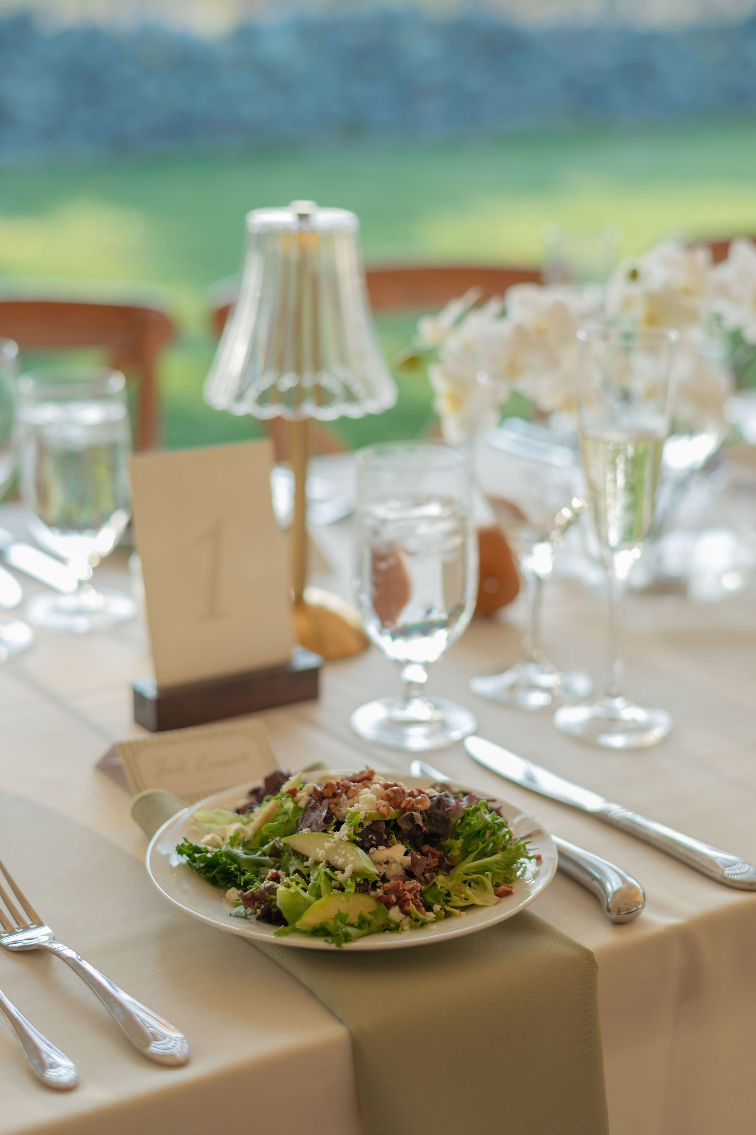 Wedding reception place setting with fresh salad, silverware, crystal glasses, small lamp, and floral arrangement on a beige tablecloth.