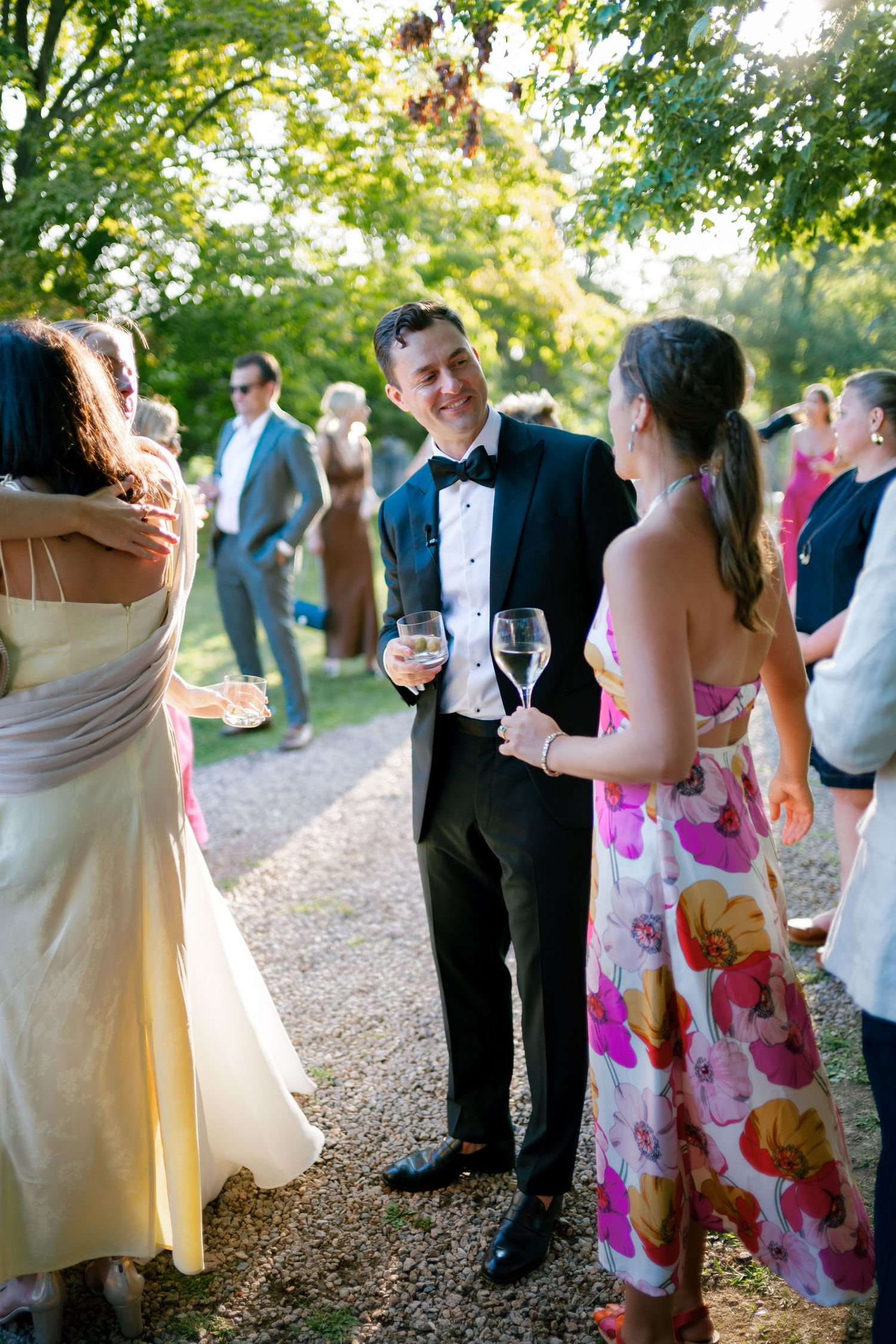 Groom in tuxedo holding champagne glasses conversing with guests at a sunny outdoor wedding reception.