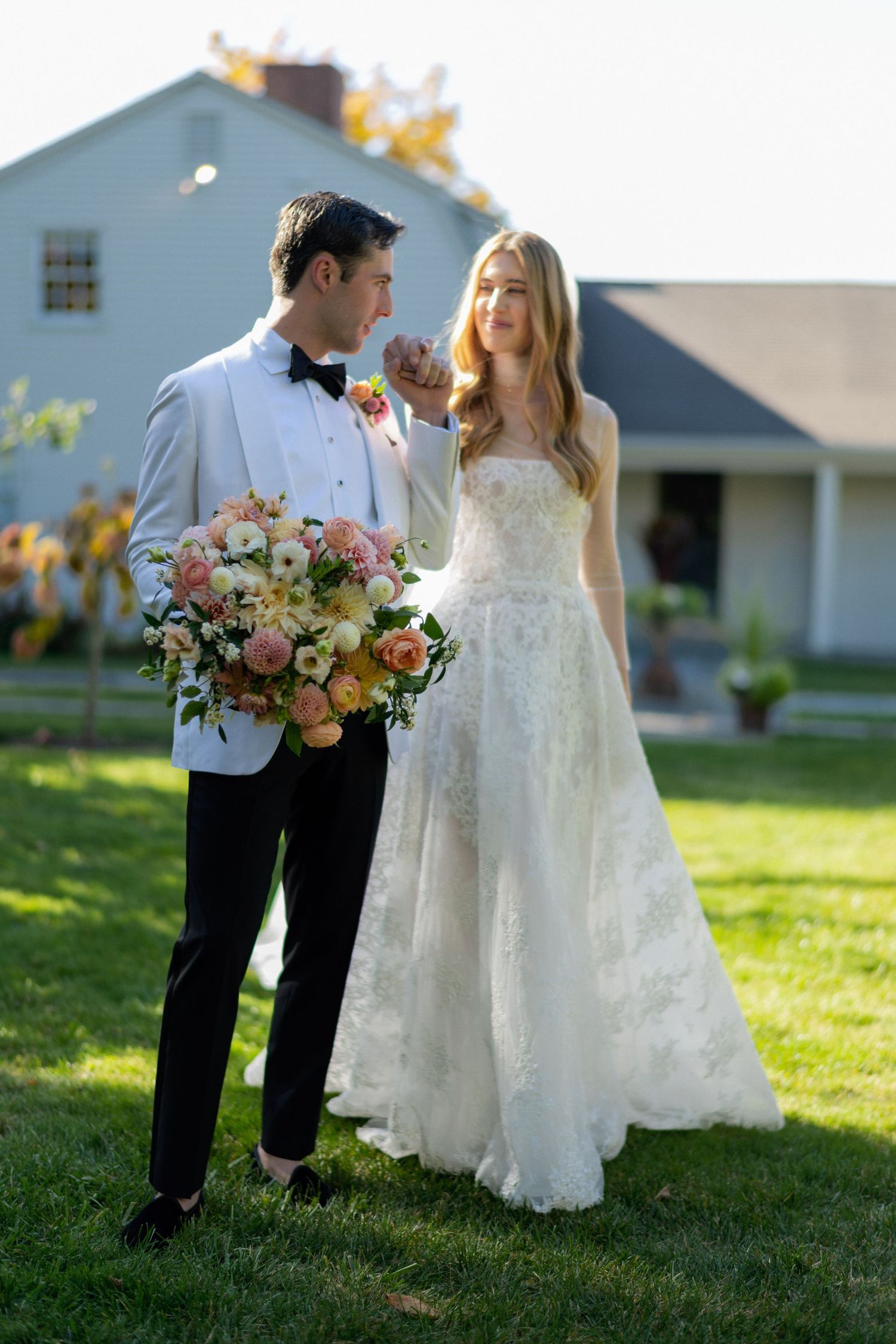 Wedding couple in formal attire posing with floral bouquet in front of farmhouse.