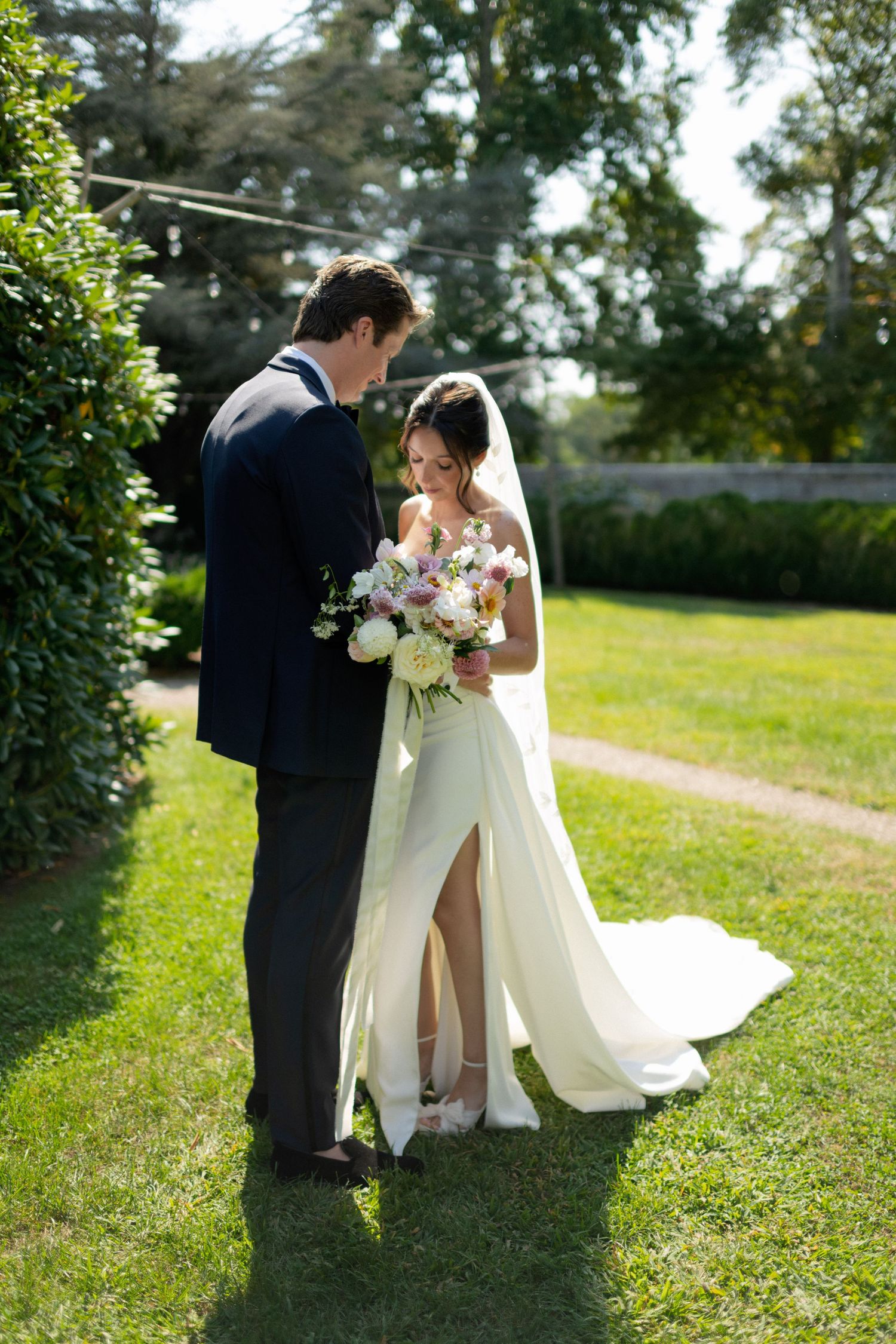 Wedding couple standing together on a sunlit garden path with elegant floral bouquet.