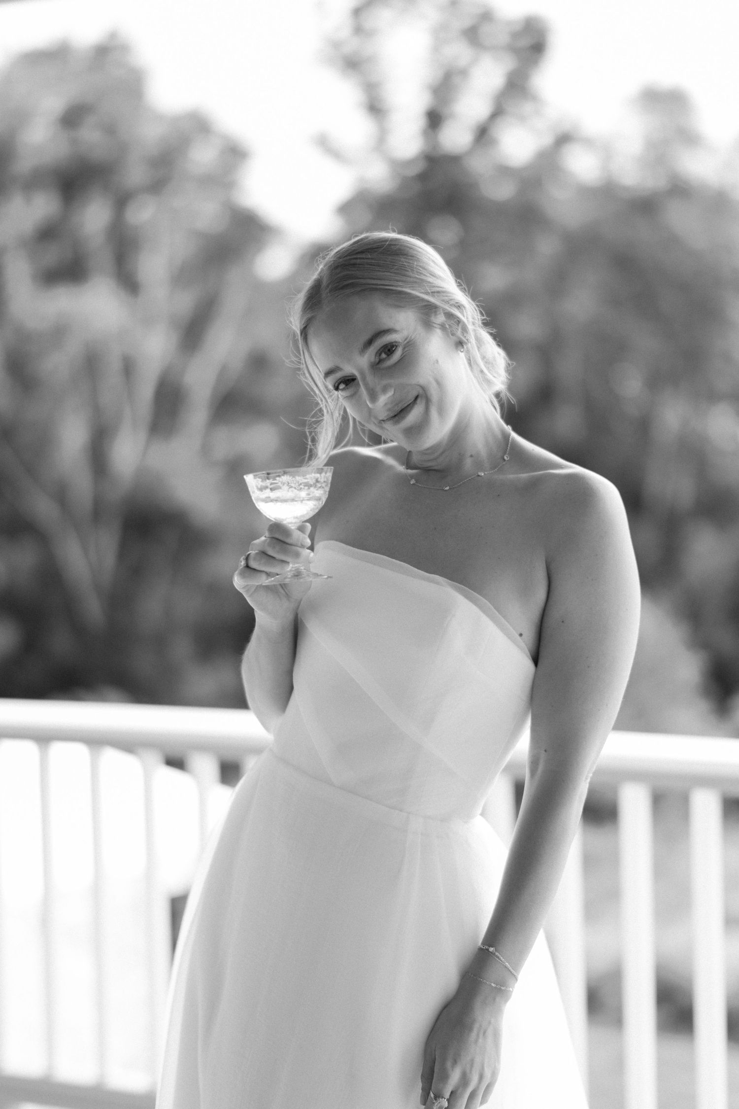 A woman in a white dress holds a champagne glass while smiling in black and white photography.