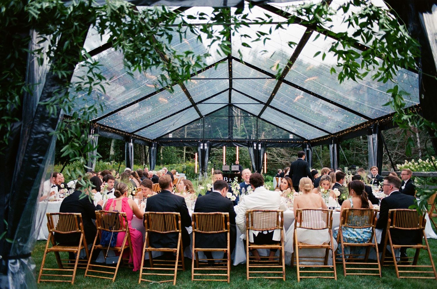 Clear tent wedding reception setup with wooden chairs and natural forest surroundings.