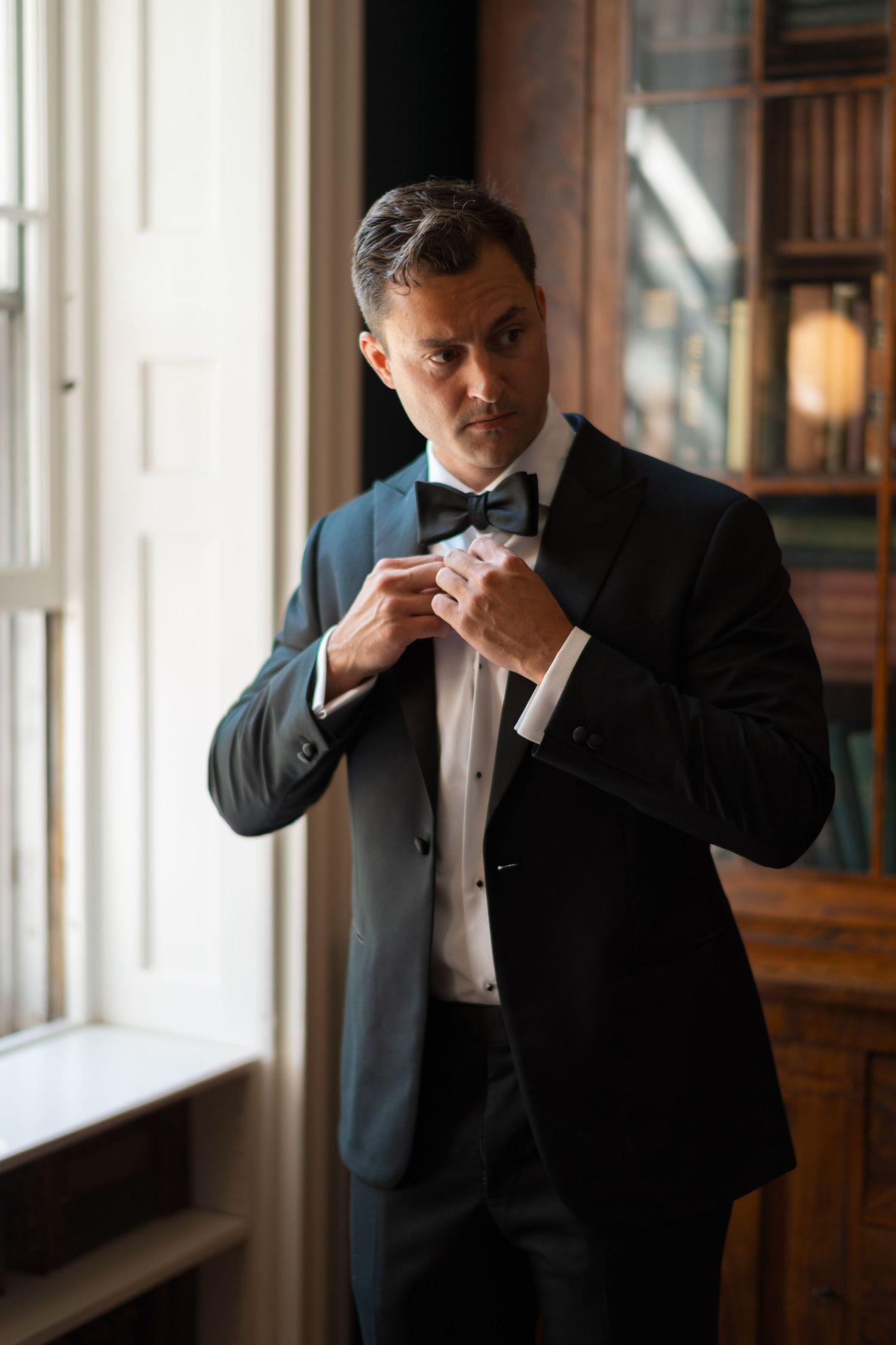 Elegant man in black tuxedo and bow tie adjusting his jacket near a window in a classic interior.