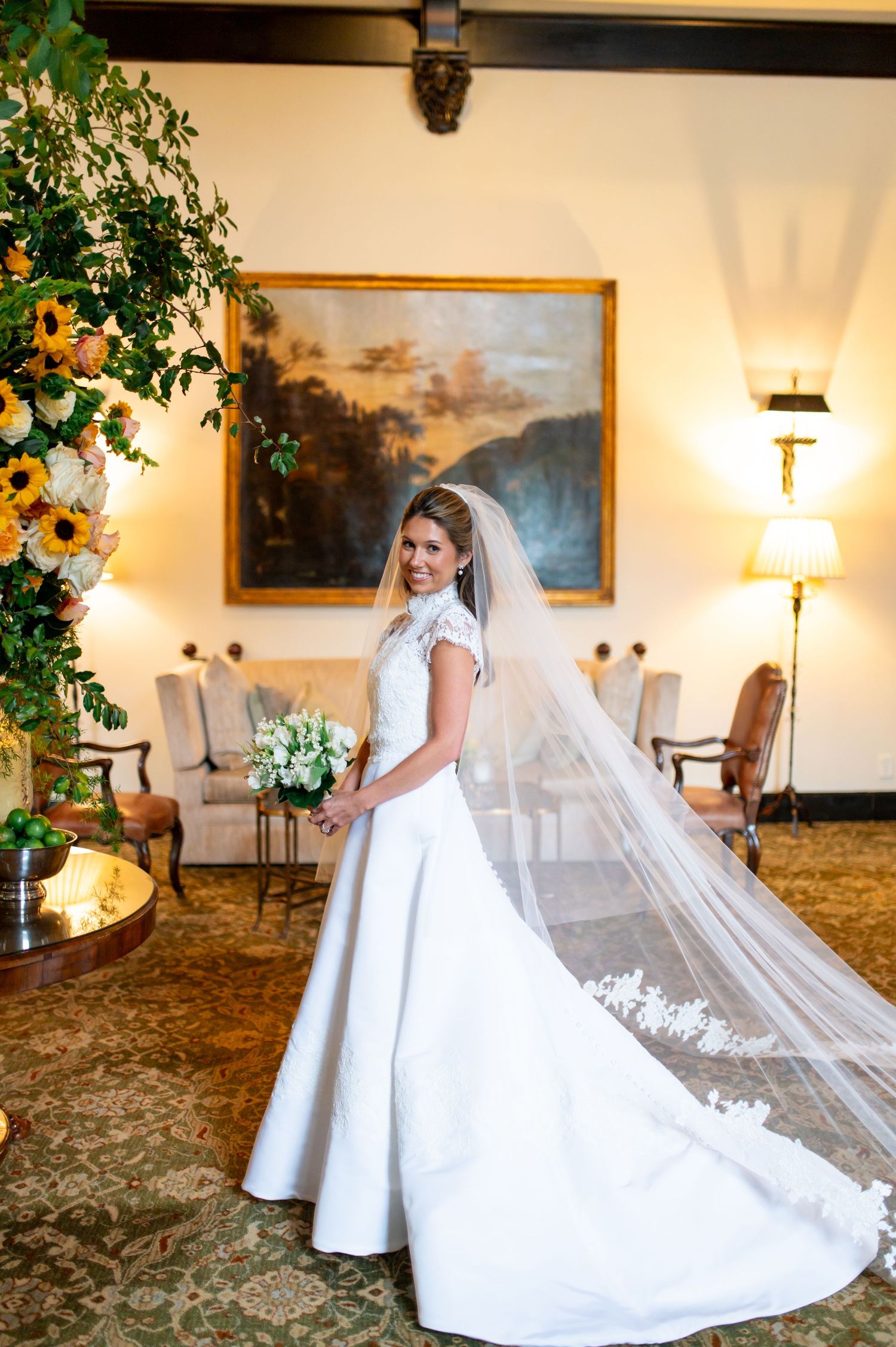 A bride in a classic white gown with long veil poses in an elegant room with landscape paintings.