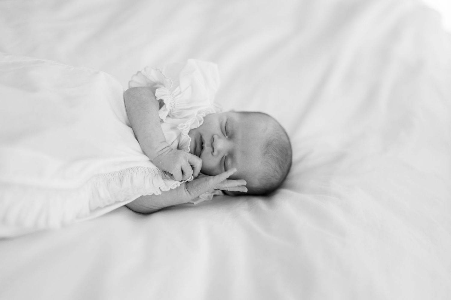 A sleeping newborn baby wearing white clothing rests peacefully on soft white bedding.