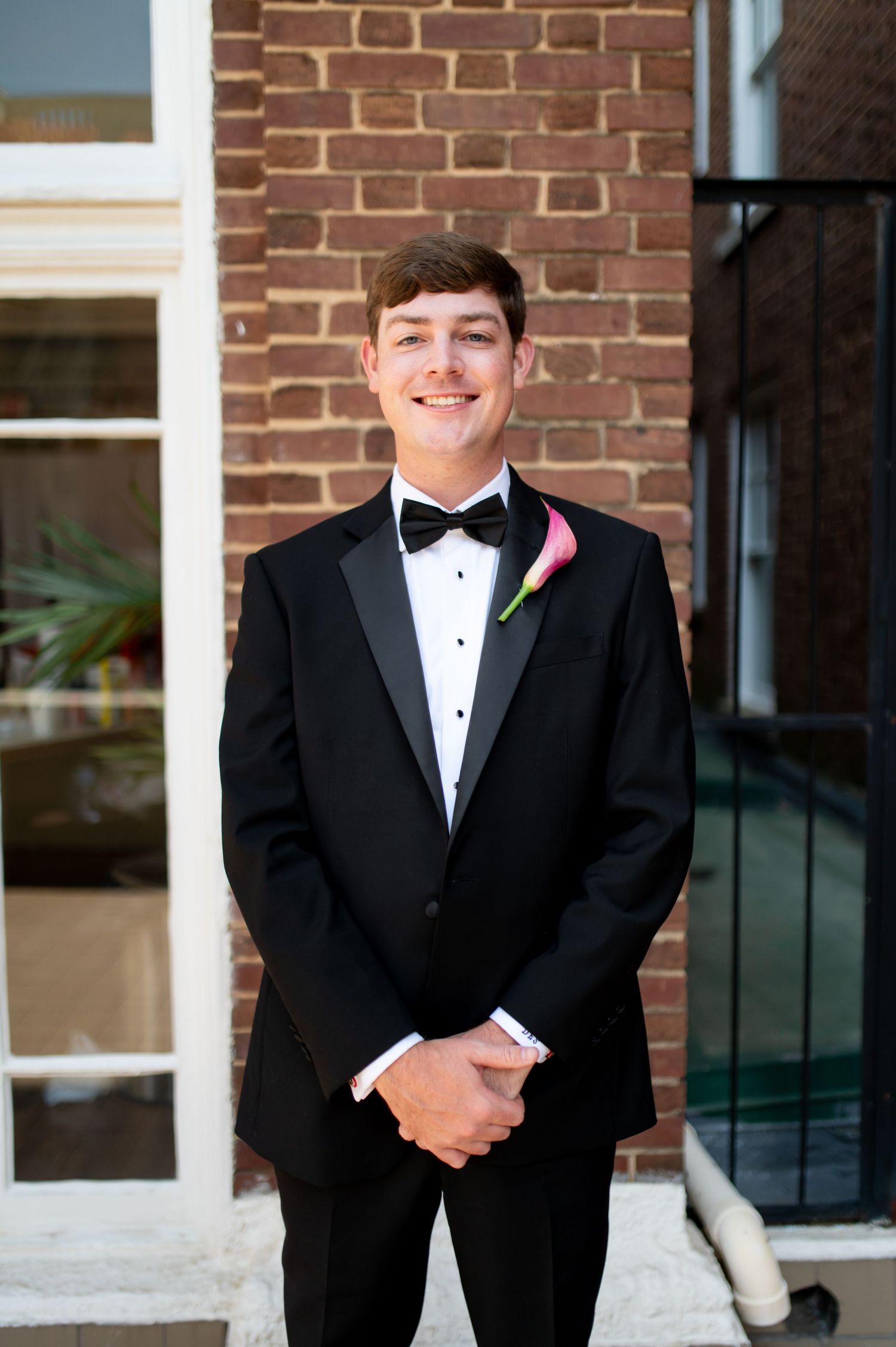 Young groomsman in black tuxedo with bow tie and pink calla lily boutonniere smiling outdoors.