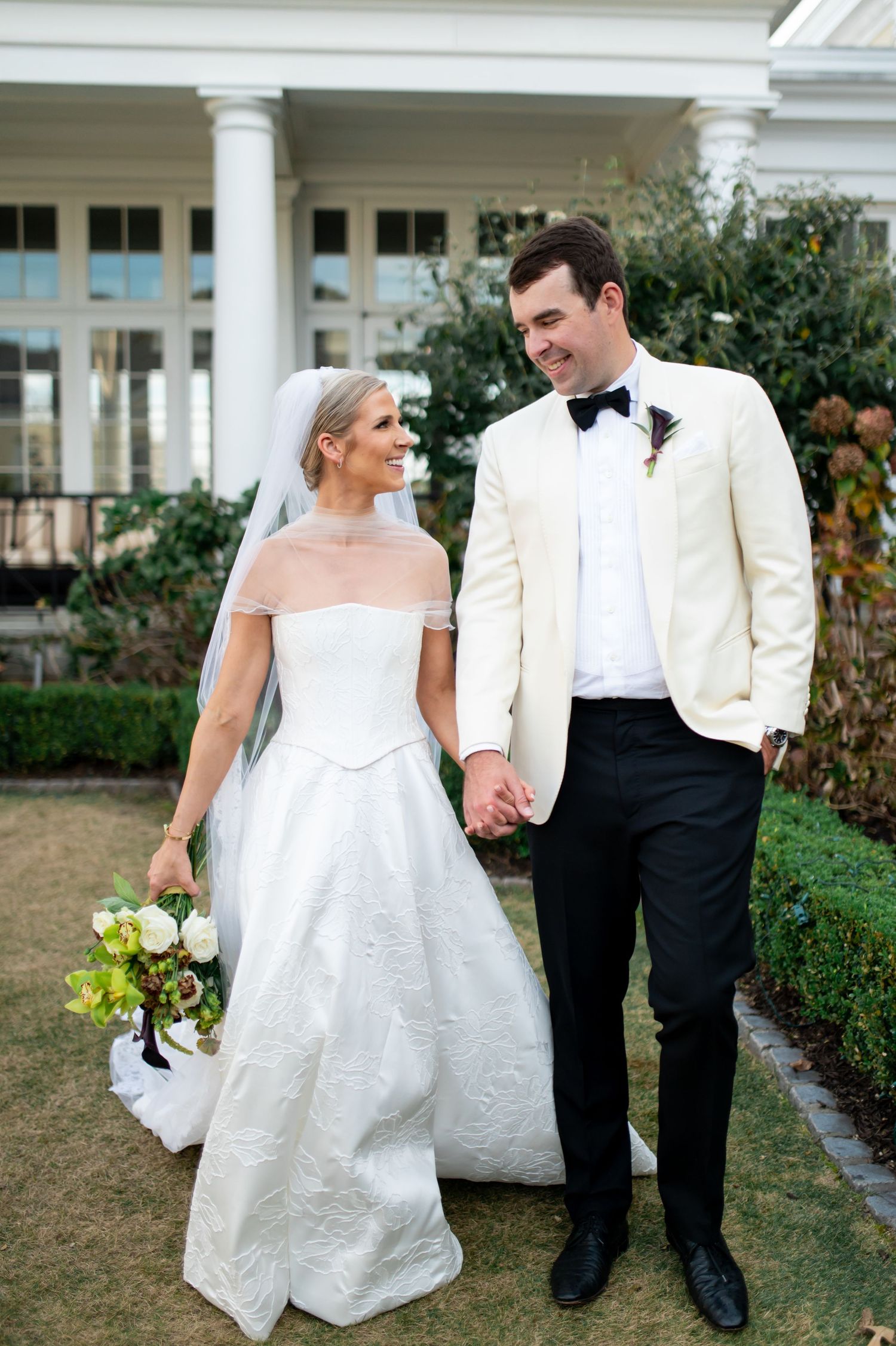 Elegant wedding couple strolling through garden grounds of a classic white columned venue.