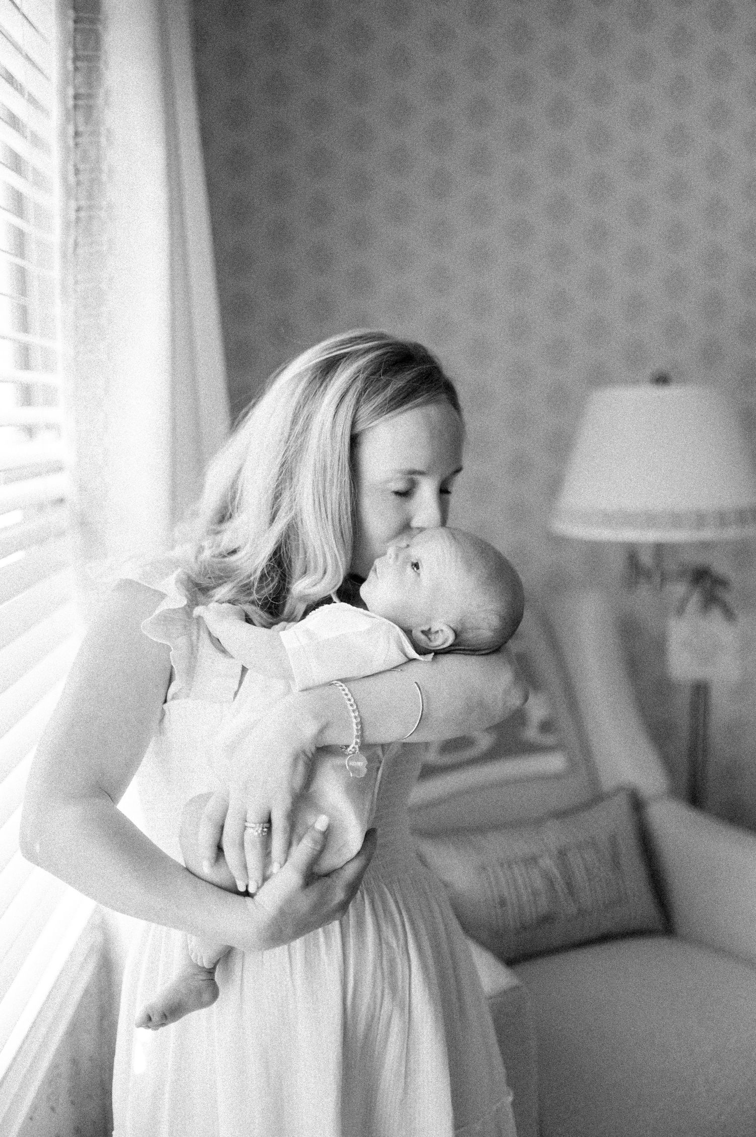 Black and white photo of a mother tenderly kissing her newborn baby near a window in a softly lit room.