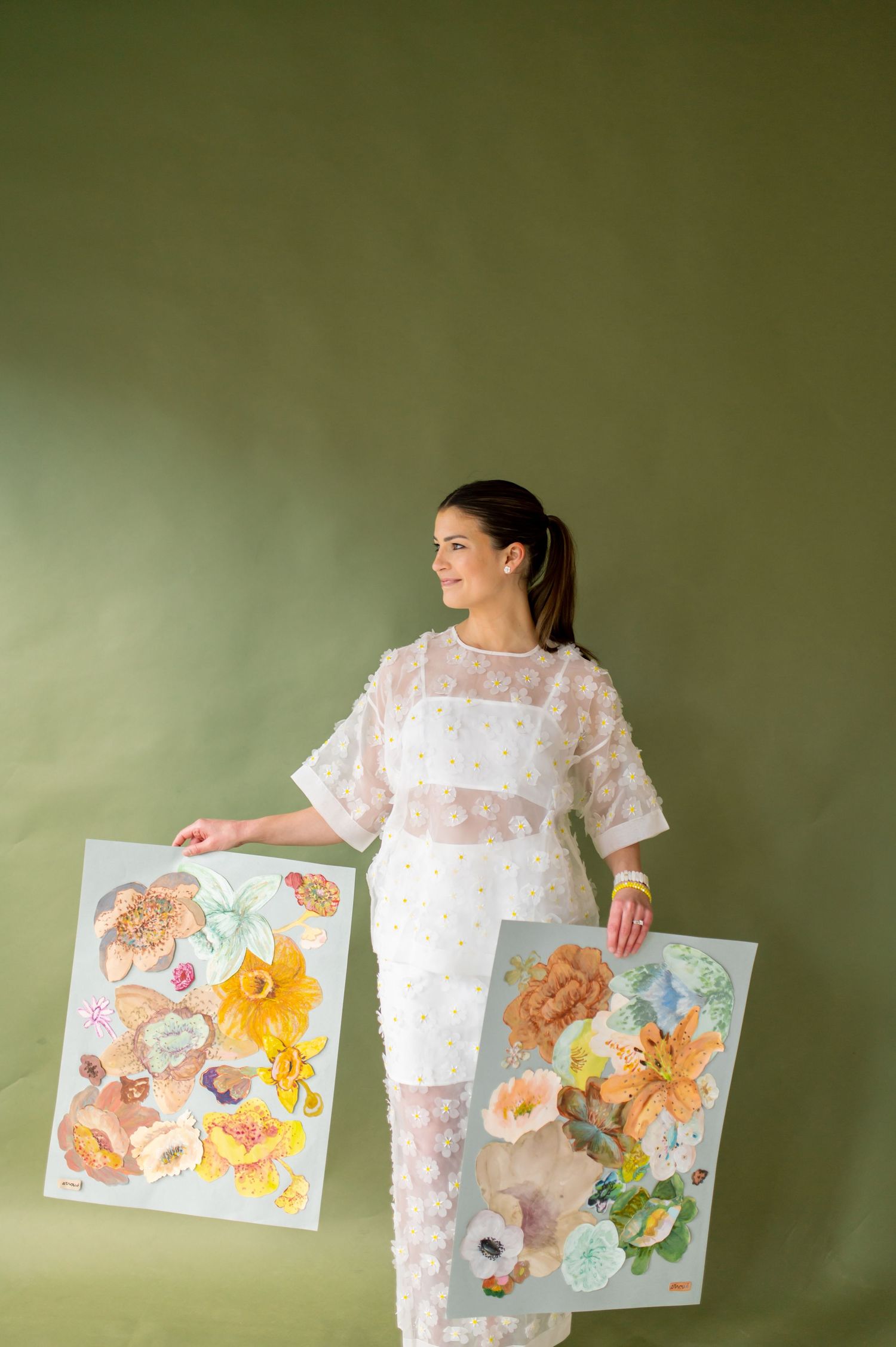 Woman in white lace dress holding two floral watercolor art prints against a green background.