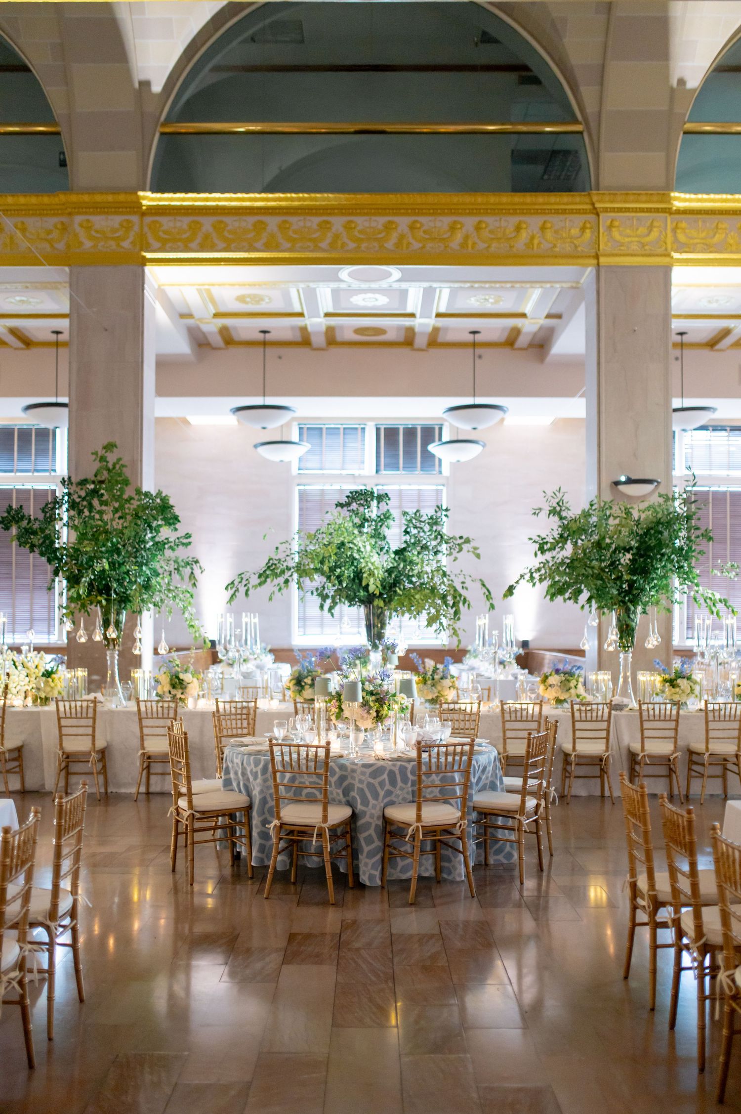 Elegant wedding reception hall with tall greenery centerpieces, gold chiavari chairs, and ornate gold ceiling details.