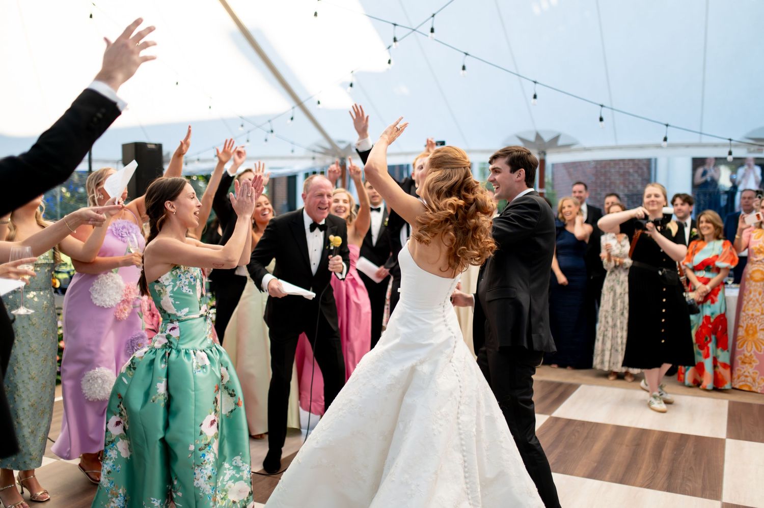 Bride and groom dance joyfully on white dance floor under tent as guests celebrate with raised hands.
