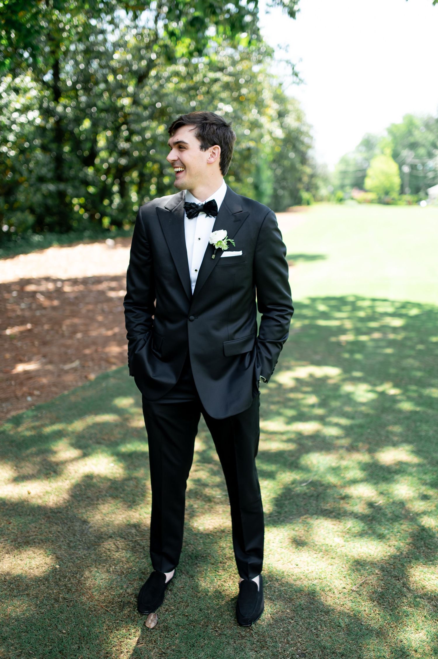 Groom in black tuxedo with bow tie and white boutonniere smiling outdoors on green lawn.