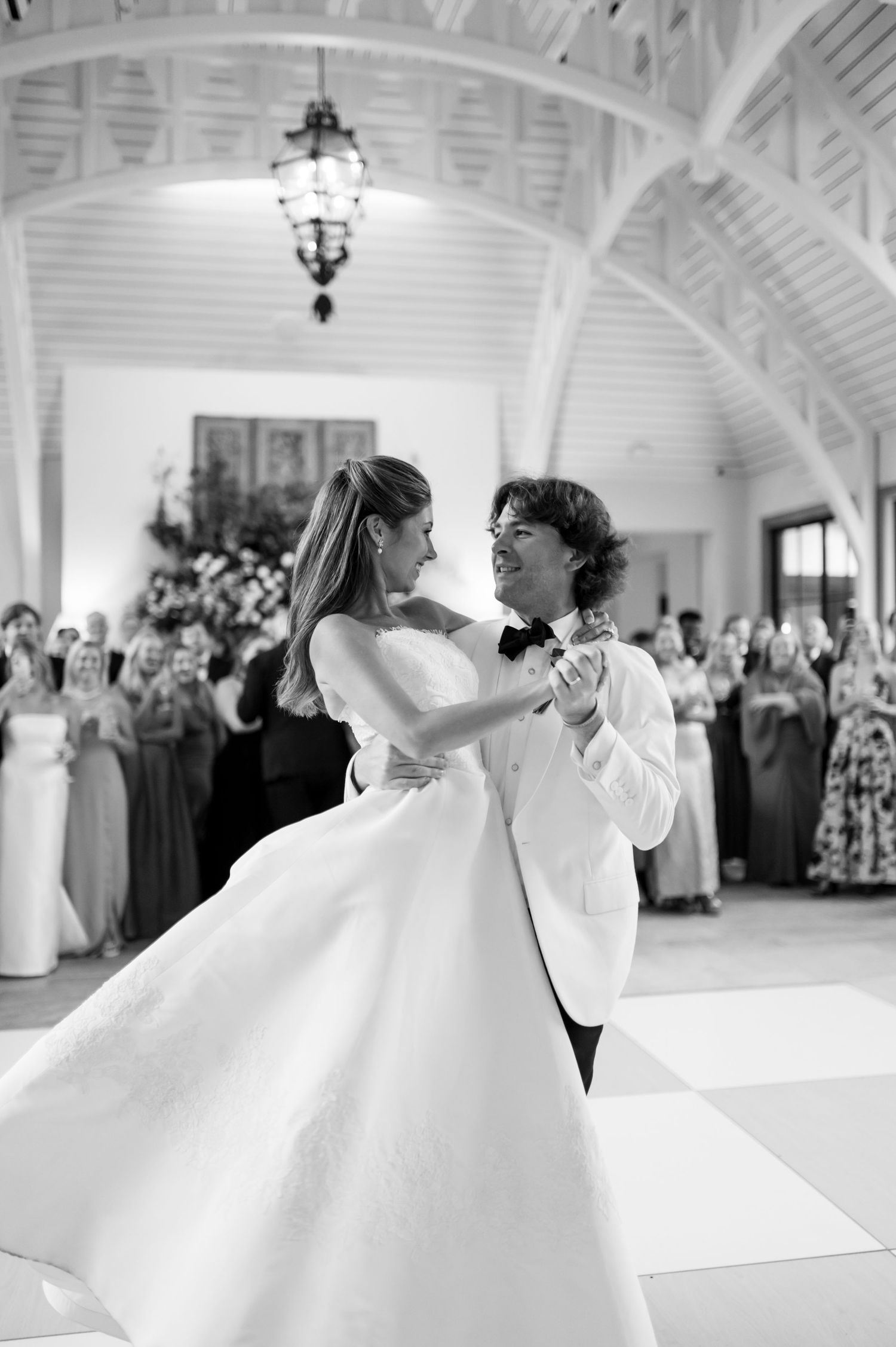 Black and white photo of couple dancing under arched ceiling with elegant chandelier.