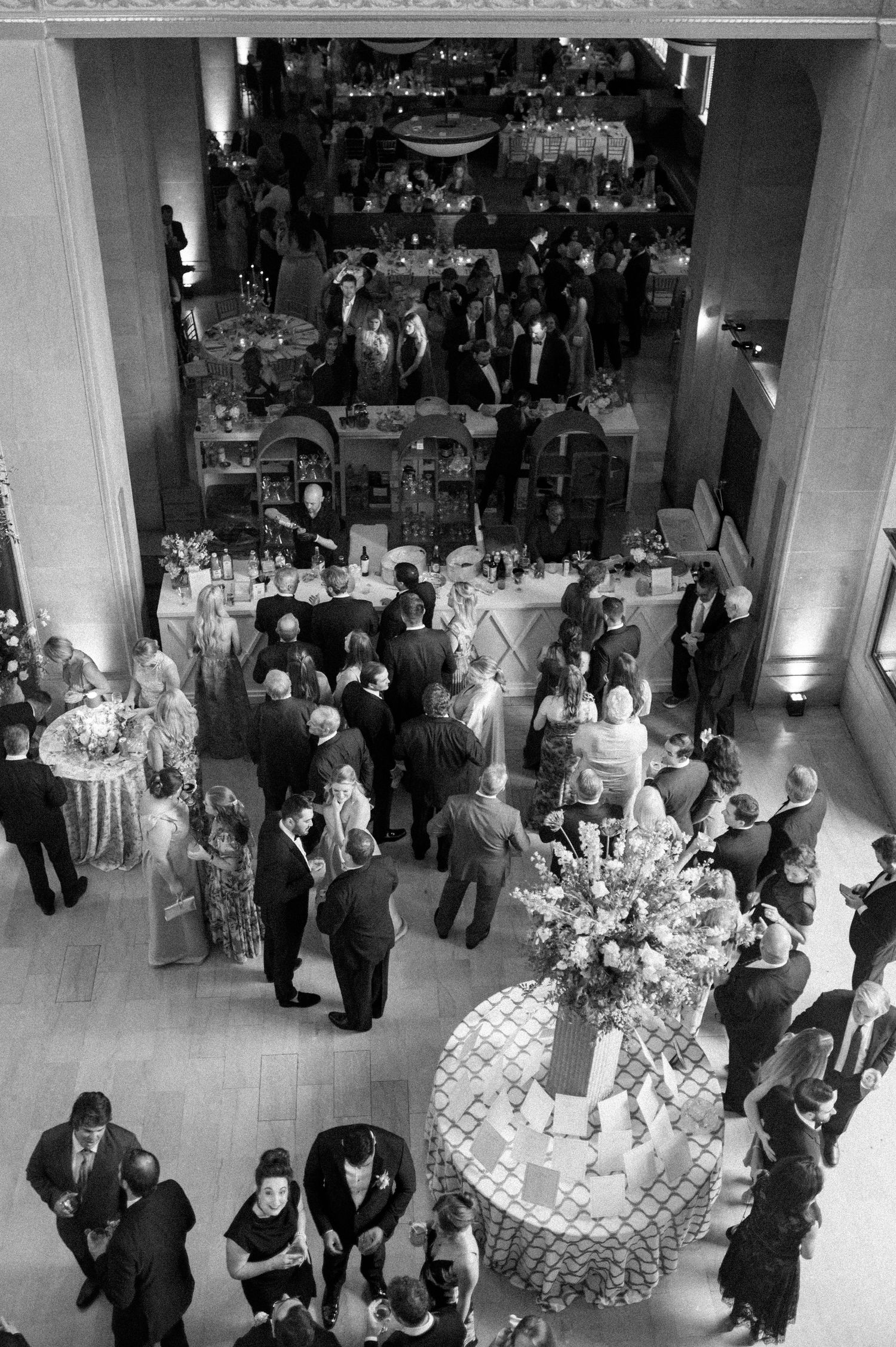 Aerial black and white photo of an elegant indoor gala event with guests mingling around decorated tables.