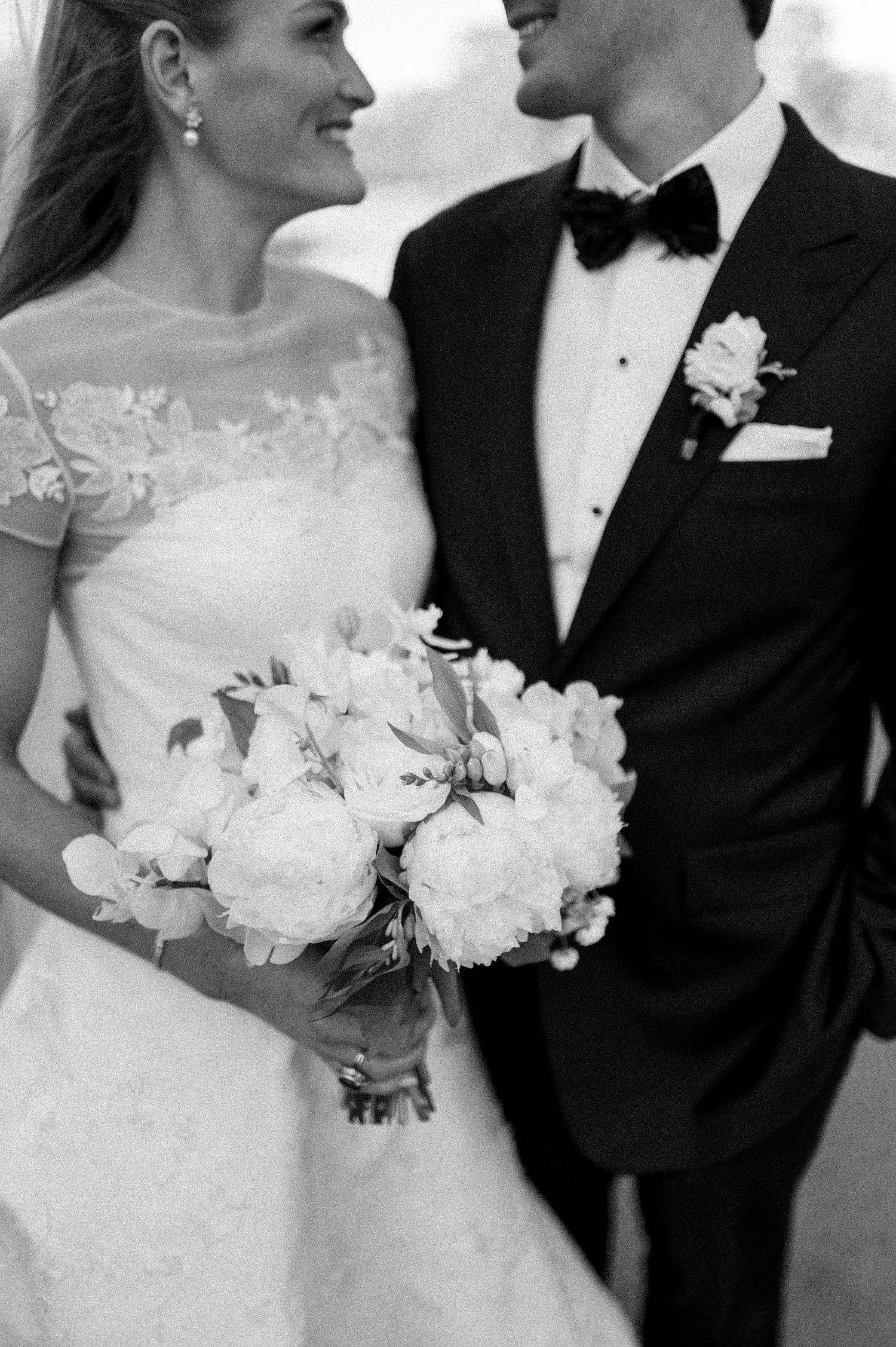 Black and white close-up of bride holding white peony bouquet beside groom in classic black tuxedo.
