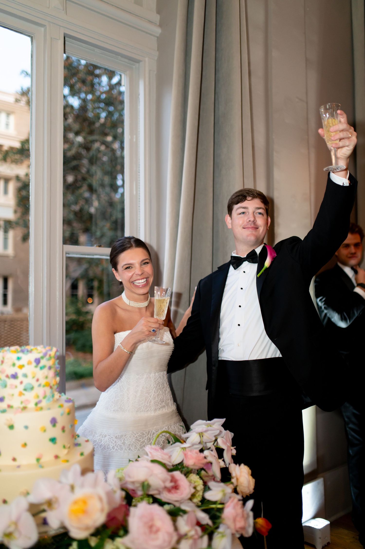 Bride and groom raise champagne glasses in toast beside floral wedding cake at elegant reception venue.
