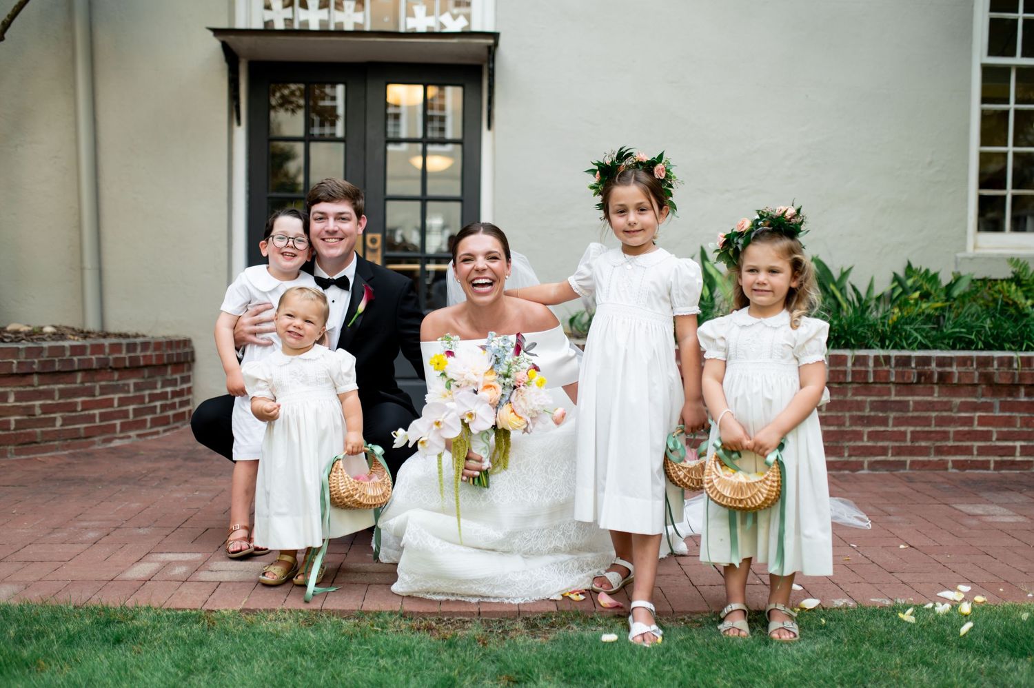 Bride and groom pose with flower girls in white dresses and floral crowns outside an elegant brick building.