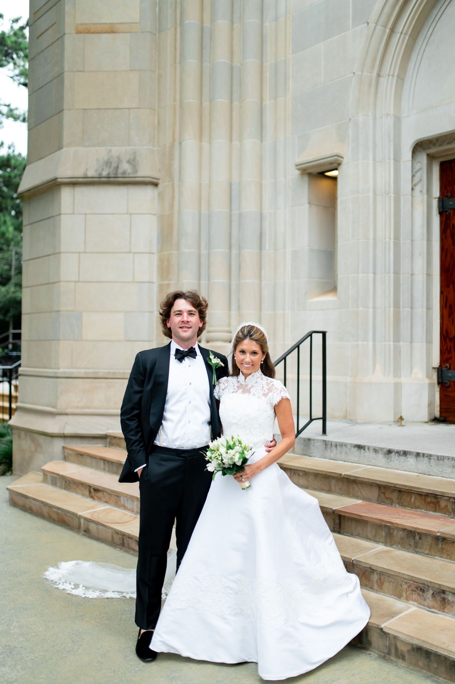 Newlyweds pose playfully on church steps in formal wedding attire.