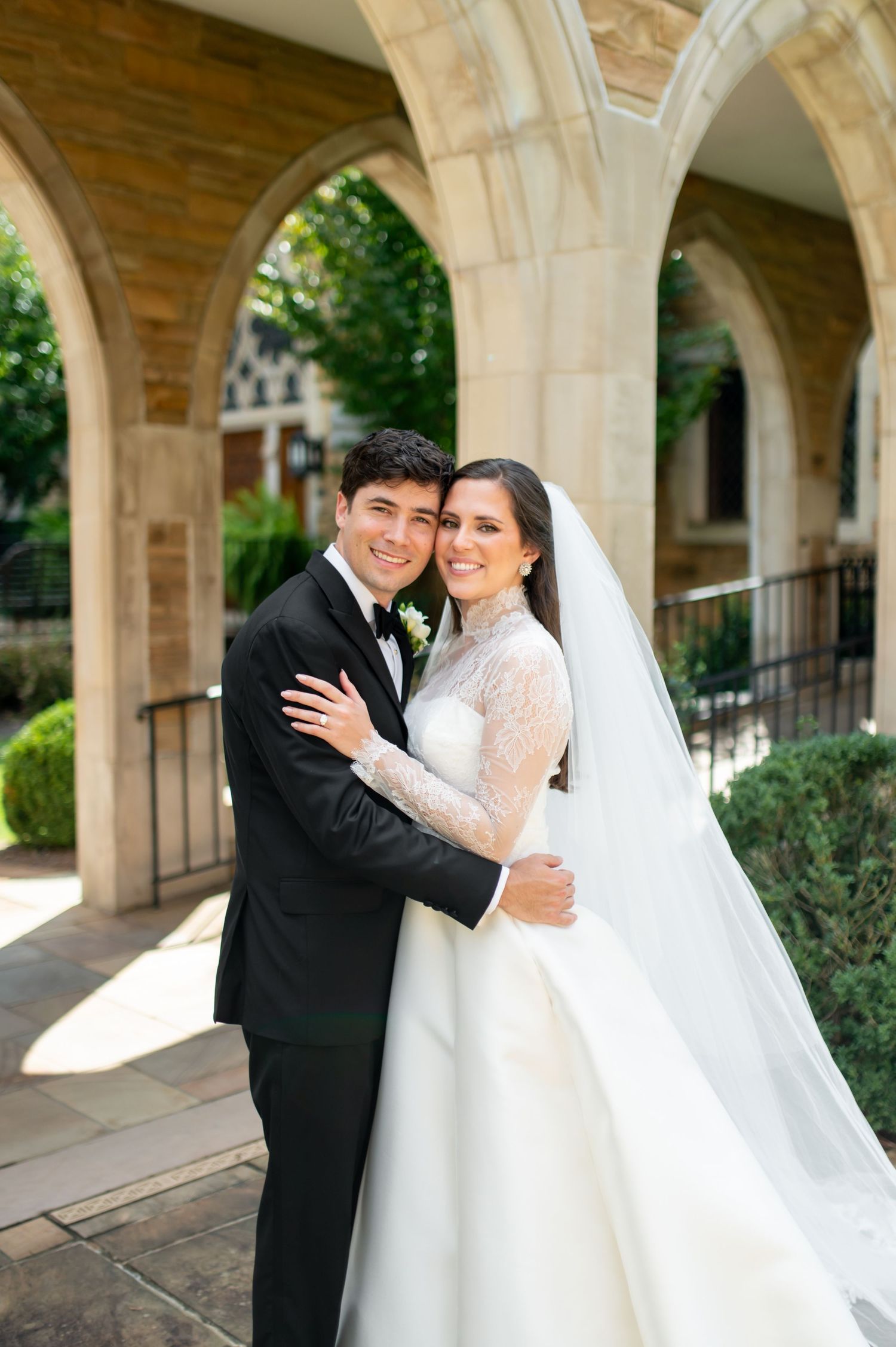 Bride and groom embrace outdoors under stone arches, wearing wedding gown with lace sleeves and classic black tuxedo.