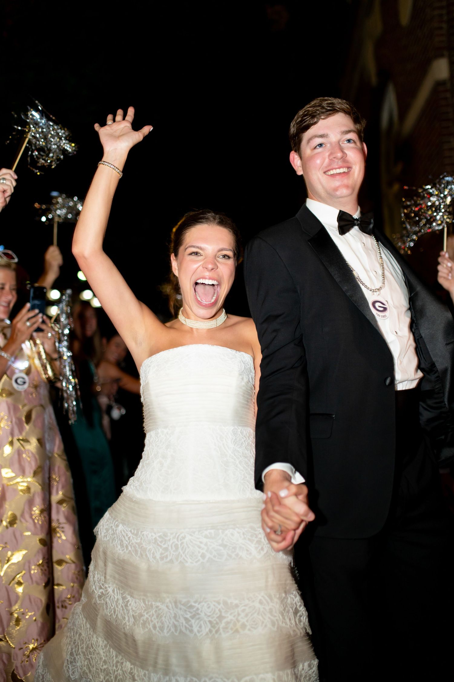 Exuberant bride raises arm during sparkler send-off exit with groom at nighttime wedding.
