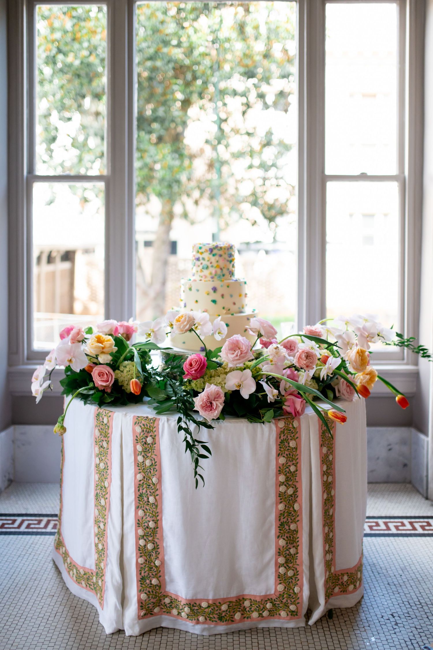 Elegant wedding cake table adorned with lush pink roses, tulips, and greenery on embroidered white tablecloth by windows.
